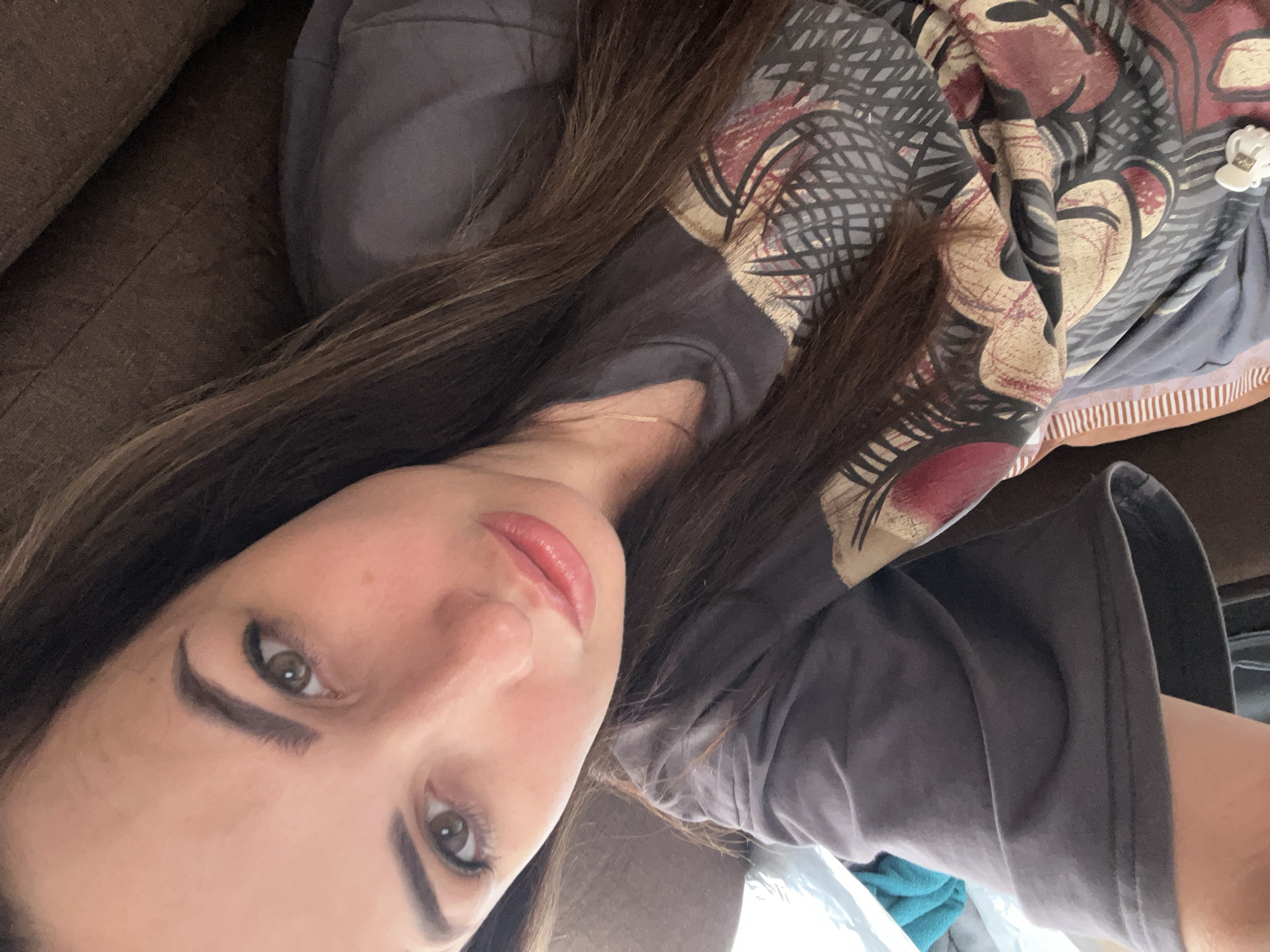 A woman is lying on a couch, taking a selfie. She has long, brown hair and brown eyes. She is wearing a gray t-shirt with a graphic print on it. The couch appears to be made of brown fabric. The lighting in the image suggests that it was taken indoors during the daytime. The background is blurred, but it appears to be a room with some blankets or pillows.