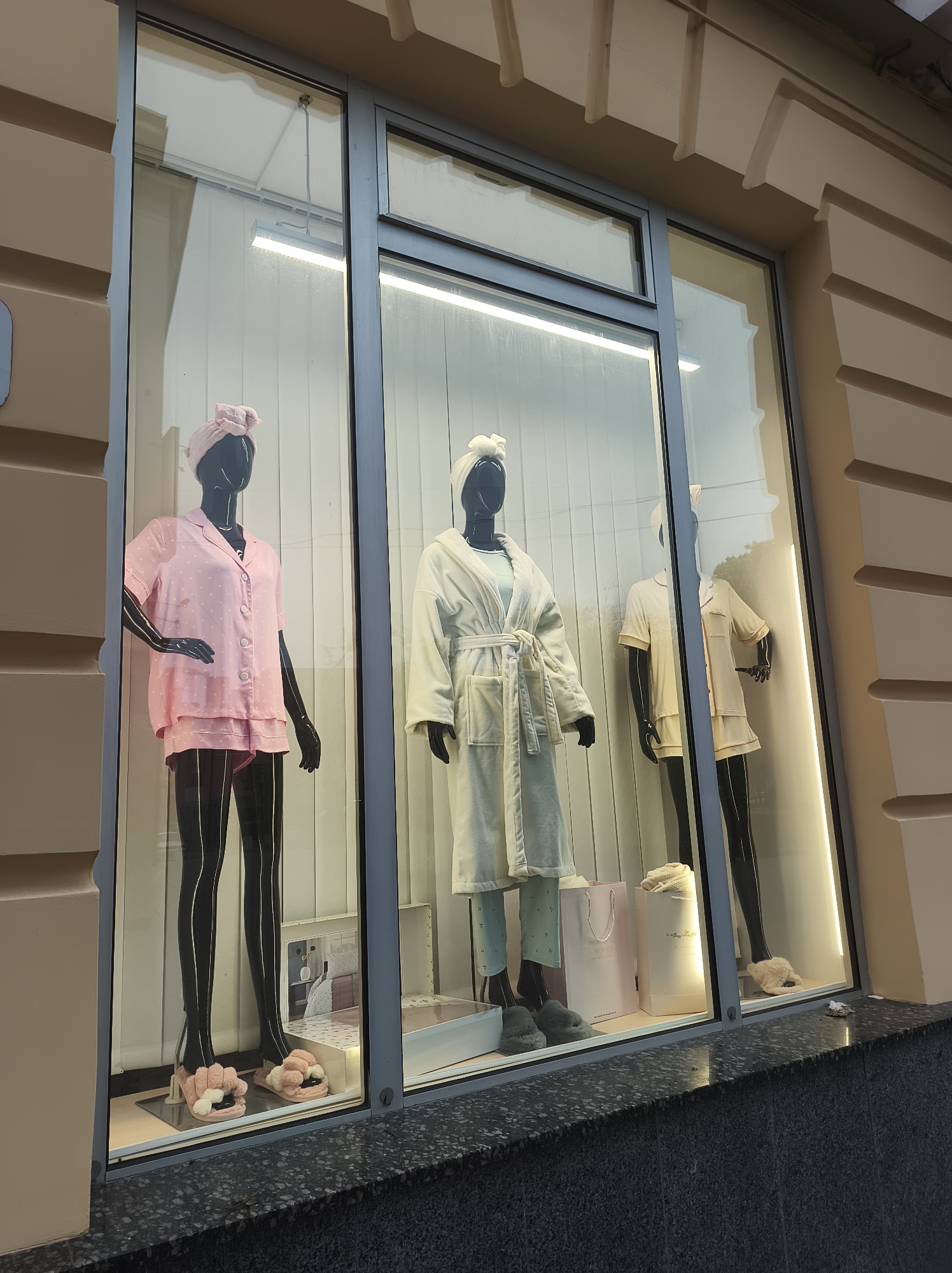 The image captures a storefront display of women's pajamas in Ternopil, Ukraine. Three mannequins with shiny black skin and hair are positioned behind glass. The mannequin on the left wears a pink, short-sleeved pajama set with white polka dots and pink slippers. The center mannequin is clad in a white bathrobe, light green pajama pants with white spots, a white head wrap and gray slippers. The mannequin on the right is wearing a tan pajama set and beige slippers. Each mannequin wears a head wrap. The window display also includes product boxes and shopping bags. The facade of the building is pale beige with horizontal architectural details. A gray granite-like ledge runs below the windows.
