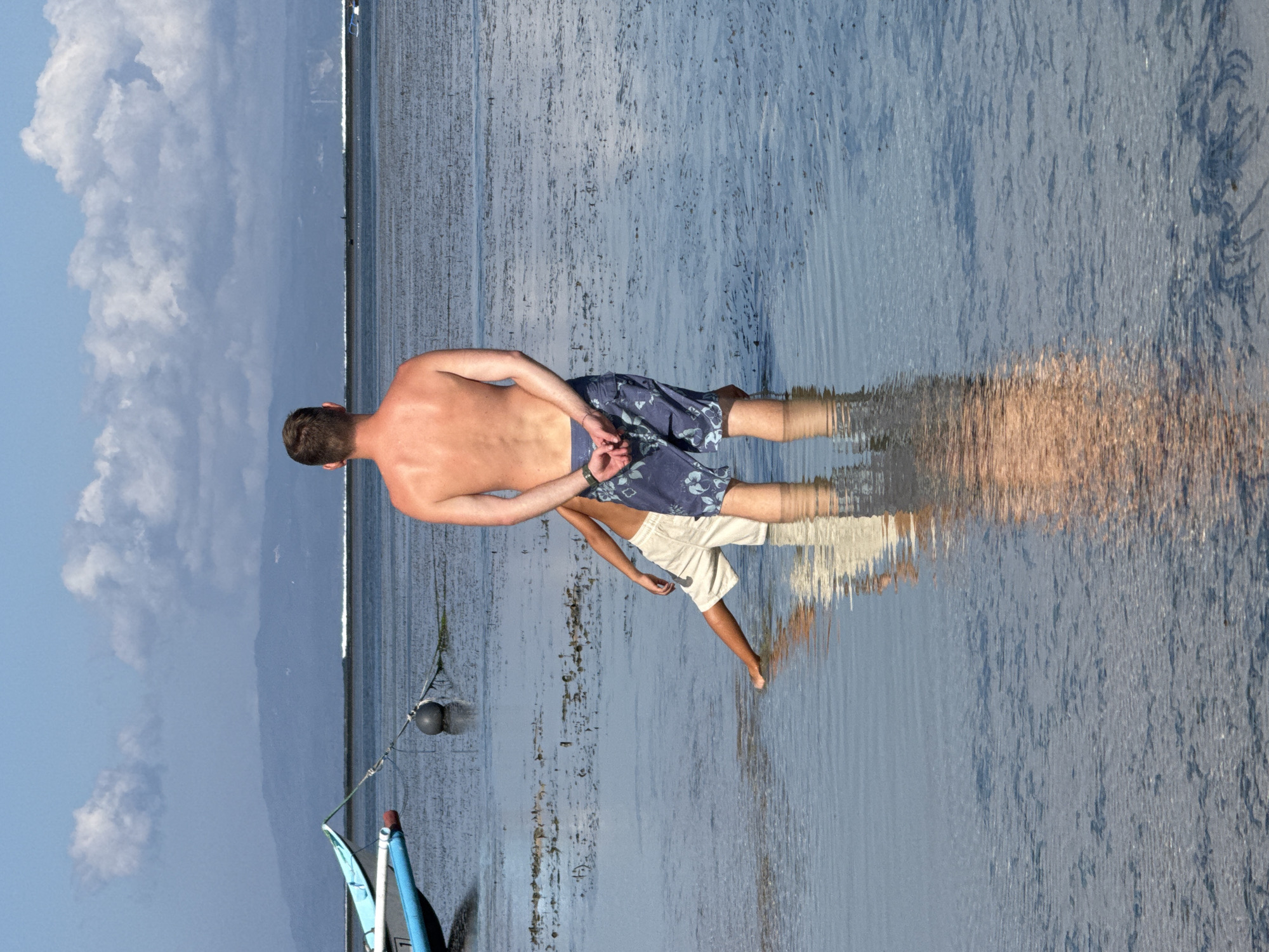 A man, facing away from the camera, stands in shallow water. He is wearing floral patterned swim trunks and has his hands clasped behind his back. To his left and slightly in front of him, another person, possibly a child, is crouched down in the water, wearing light-colored shorts. The background features a calm body of water with two boats moored in the distance. A hazy mountain range is visible on the horizon under a partly cloudy sky. The scene appears to be a beach or coastal area in Denpasar, Indonesia, likely during low tide, given the exposed seabed.