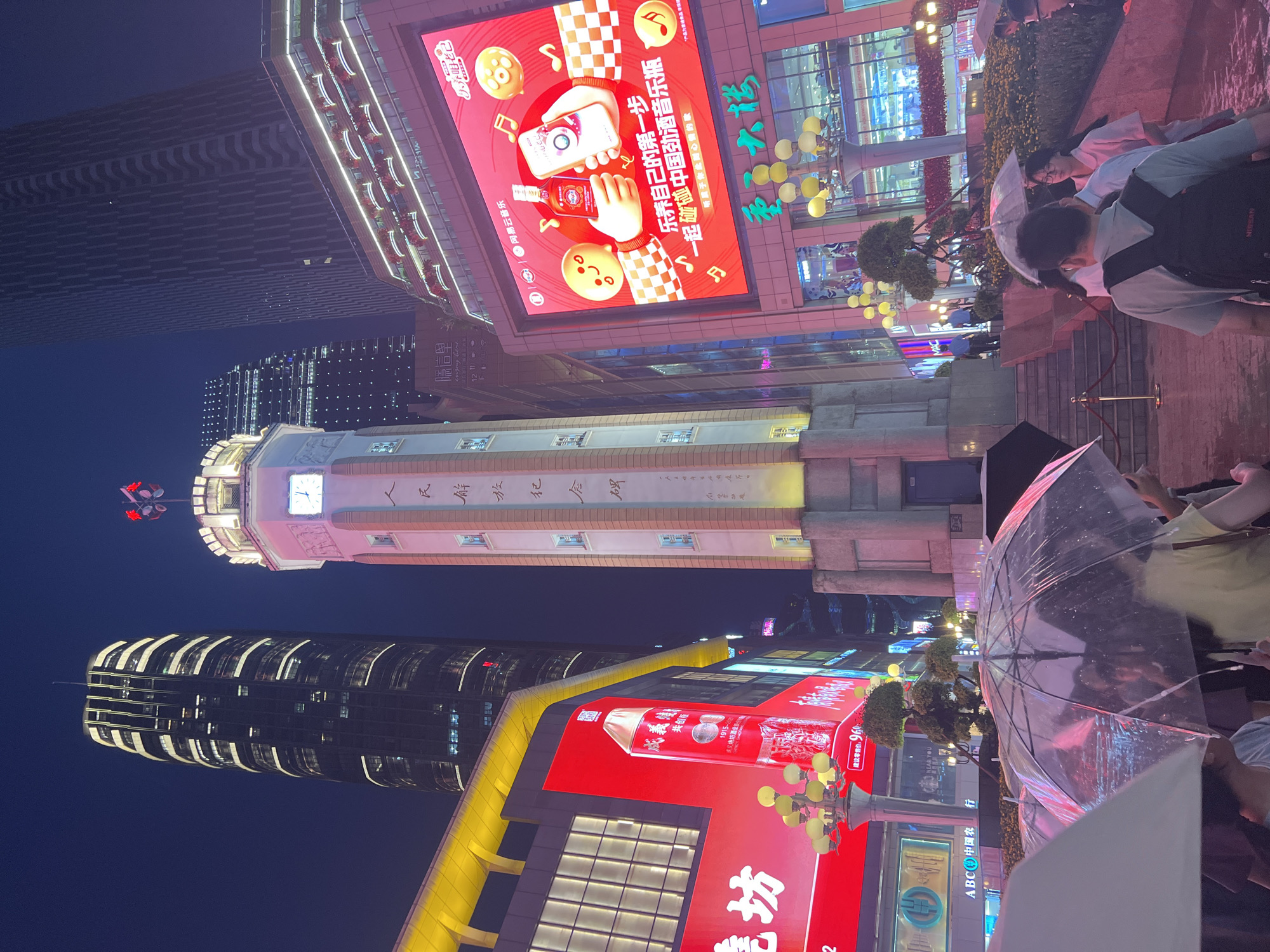 The image captures a nighttime street scene in Yuzhong District, China, characterized by vibrant neon lights and towering modern architecture. A prominent clock tower stands centrally, adorned with Chinese characters. The foreground shows several individuals holding umbrellas, suggesting it might be raining or the ground is wet. Surrounding the clock tower are large illuminated billboards displaying advertisements. In the background, several tall skyscrapers dominate the skyline, their windows lit up. The overall atmosphere is one of a bustling, modern urban environment at night.