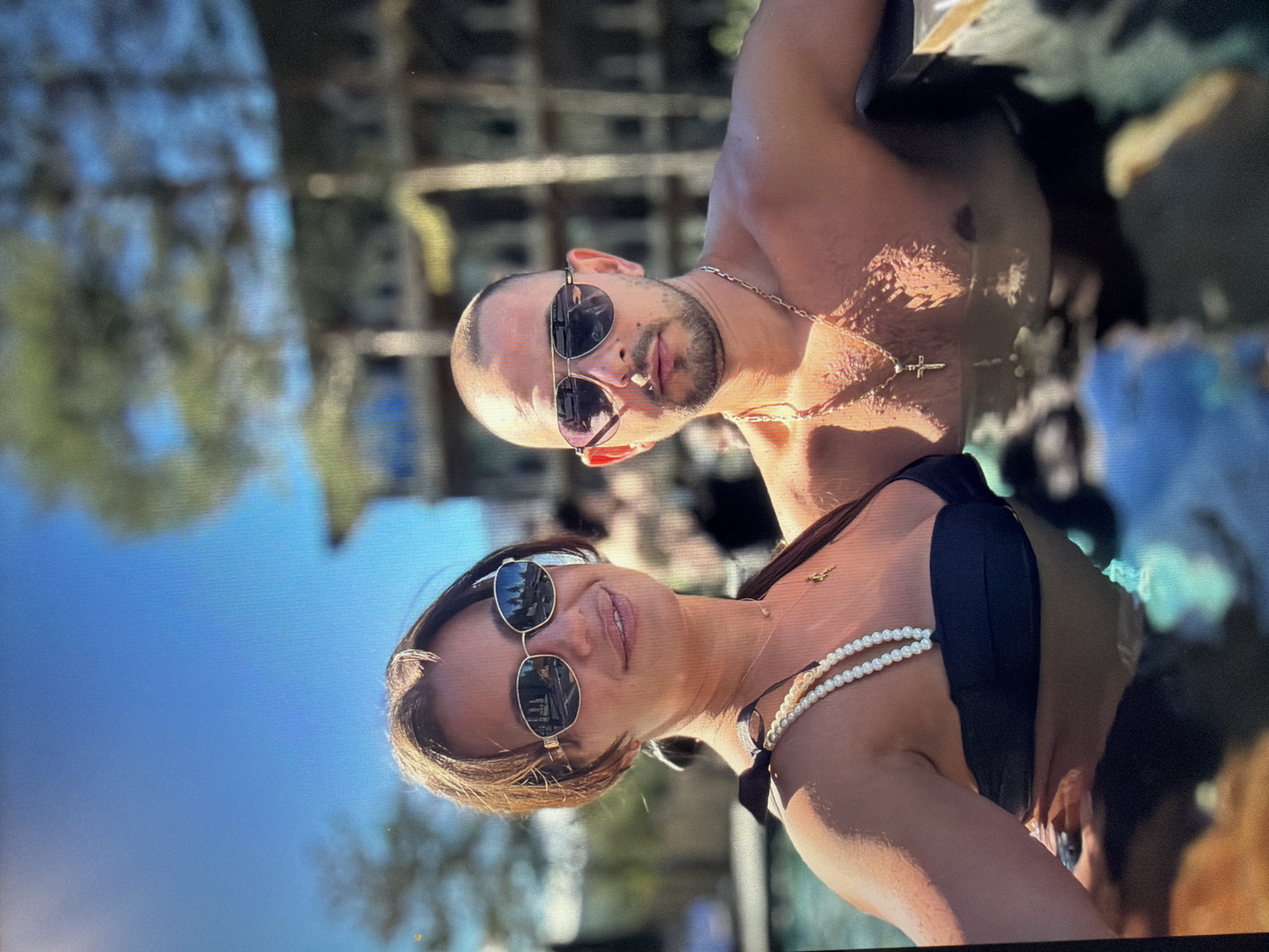A man and a woman pose closely in a pool. The man is bald with a beard and sunglasses, wearing a gold chain with a cross pendant. The woman has sunglasses and a pearl necklace and is wearing a black swimsuit. The location appears to be a sunny outdoor area in the city of Burgas, Bulgaria, with visible tree reflections in their sunglasses and distant building structures in the background. They are both looking at the camera.