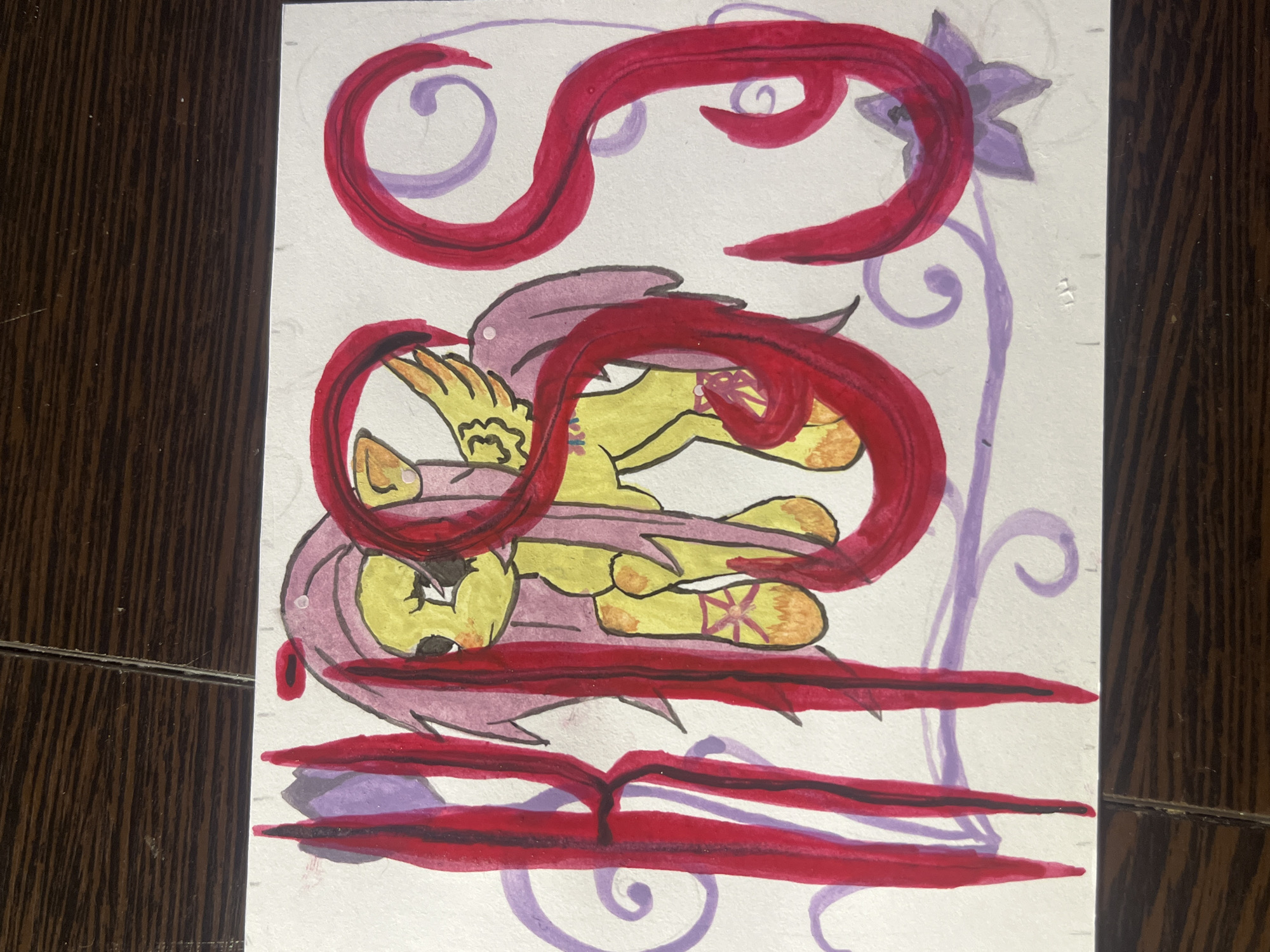 The image is a drawing on white paper. The main subject is a yellow pony with pink hair and wings, lying horizontally. A thick, red ribbon-like element is wrapped around the pony's body. Above the pony is another red ribbon forming loops. In the background, a light purple vine with a flower at the top right is visible. The artwork is placed on a dark wooden surface. There is no indication of the location being Saint Petersburg, Russia.