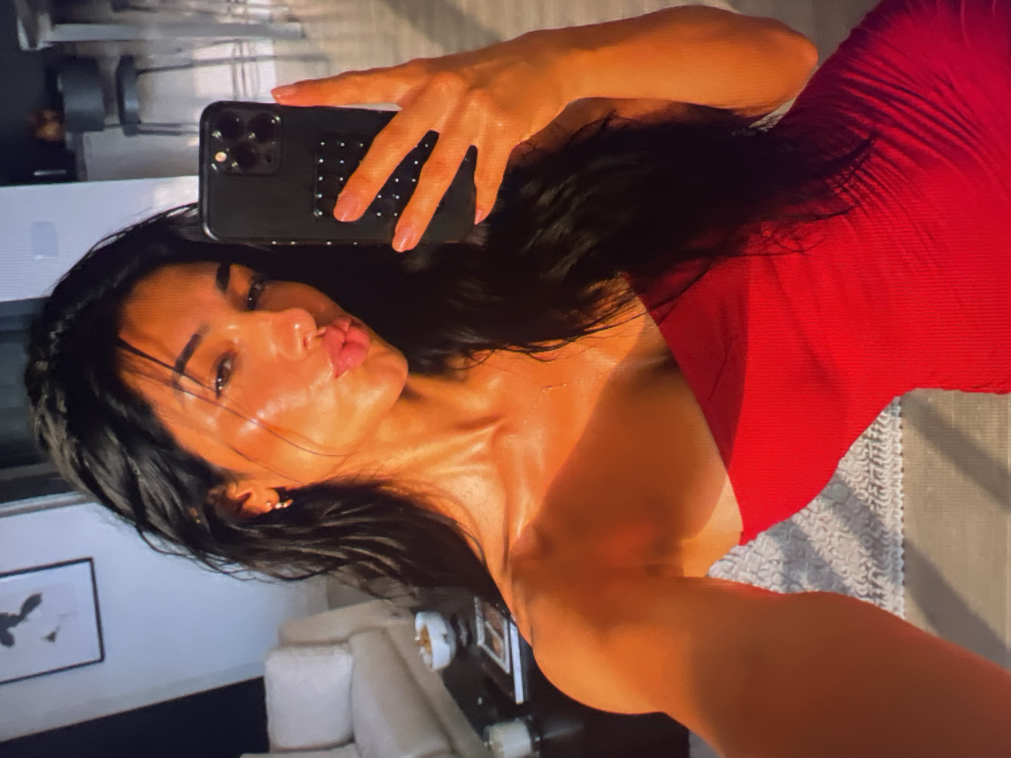 A woman takes a selfie with her phone in a dimly lit room. She is wearing a strapless red dress and her long, dark hair is loose around her shoulders. She is puckering her lips for the camera, and her eyes are slightly closed. The phone she is holding is black with a patterned case. In the background, a couch and a framed picture on the wall are visible. The overall mood of the image is playful and intimate. The lighting suggests it is nighttime or the room is otherwise dark.