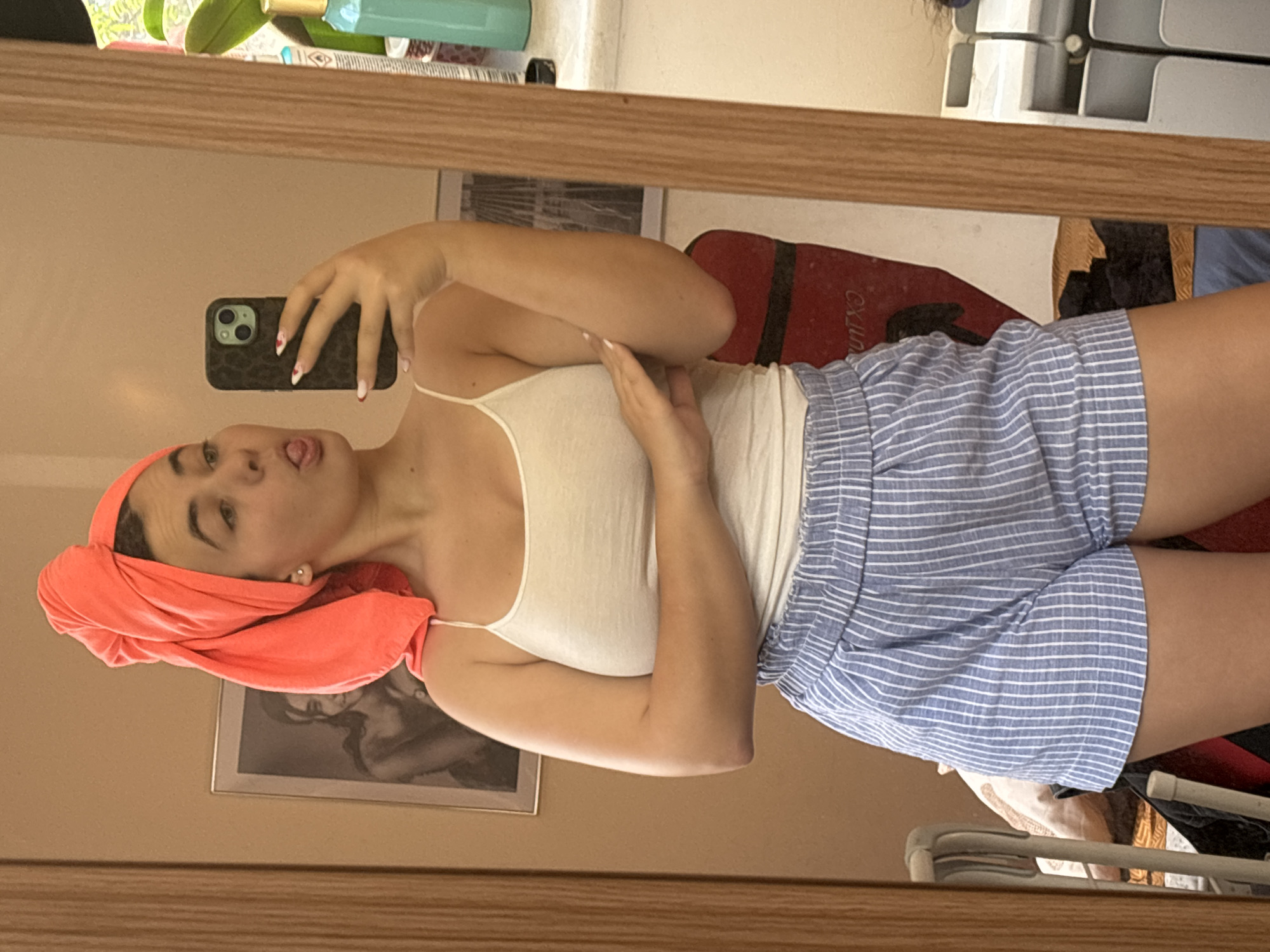 A woman in a white tank top and blue and white striped shorts stands in front of a mirror, taking a selfie with a phone in her left hand. She has an orange towel wrapped around her head. She is sticking her tongue out. The background includes a framed picture, a red item with "CXJINA" partially visible, and miscellaneous items on a shelf. The location is likely indoors, but the city of Plovdiv, Bulgaria cannot be determined from the image.