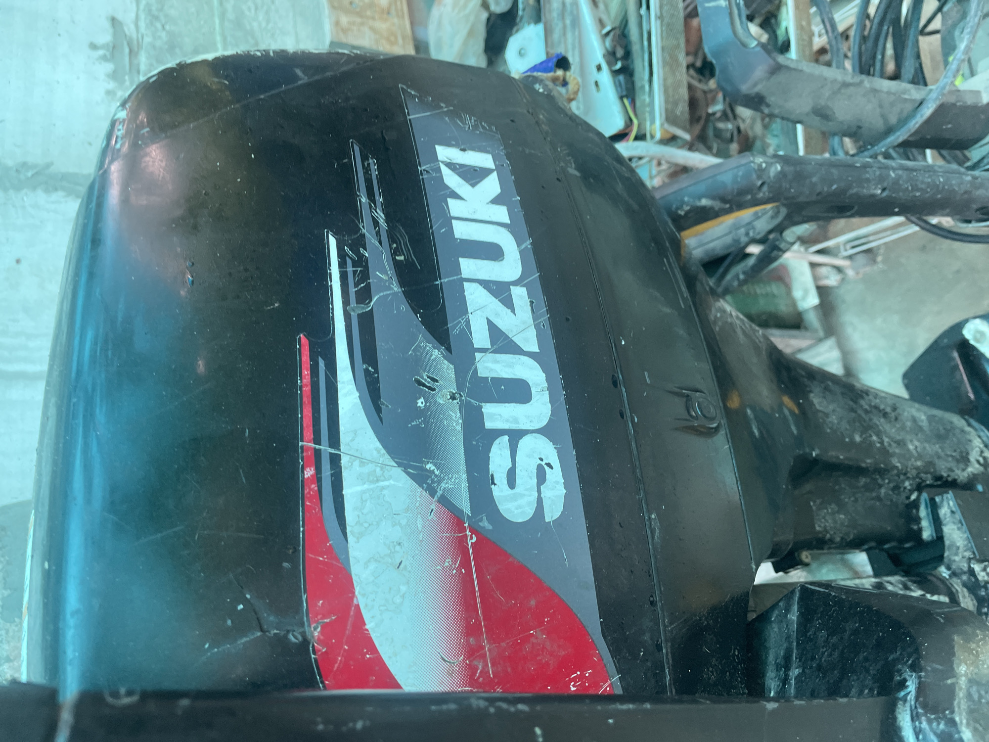 The image depicts a partially visible black Suzuki outboard motor, with a prominent Suzuki logo in white and a red and white graphic design. The motor appears used and scratched. The surrounding environment shows other mechanical parts and miscellaneous items, suggesting a garage or workshop setting. The location of the scene is not discernible from the image.