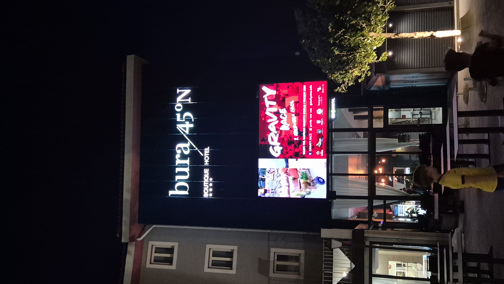 An outdoor nighttime shot captures a building facade adorned with illuminated signage. At the top, a white neon sign reads "bura 45°N BOUTIQUE HOTEL" with four stars beneath it. Below this, a large LED screen displays a vibrant advertisement for "GRAVITY RACE 1. KOLUBOVA 2024". The screen features images of people participating in what appears to be a race, with red and white as dominant colors. The building has a modern aesthetic with multiple windows and a glass-fronted entrance. A young boy wearing a yellow shirt stands in the foreground, facing left and slightly away from the camera. Another person, a child, is partially visible in the lower right corner, facing away from the camera. Outdoor tables and chairs are arranged in front of the building, suggesting an outdoor seating area for a restaurant or cafe. A tree with illuminated lights stands to the right of the building. The overall scene is well-lit by the signage and the ambient lighting of the establishment.