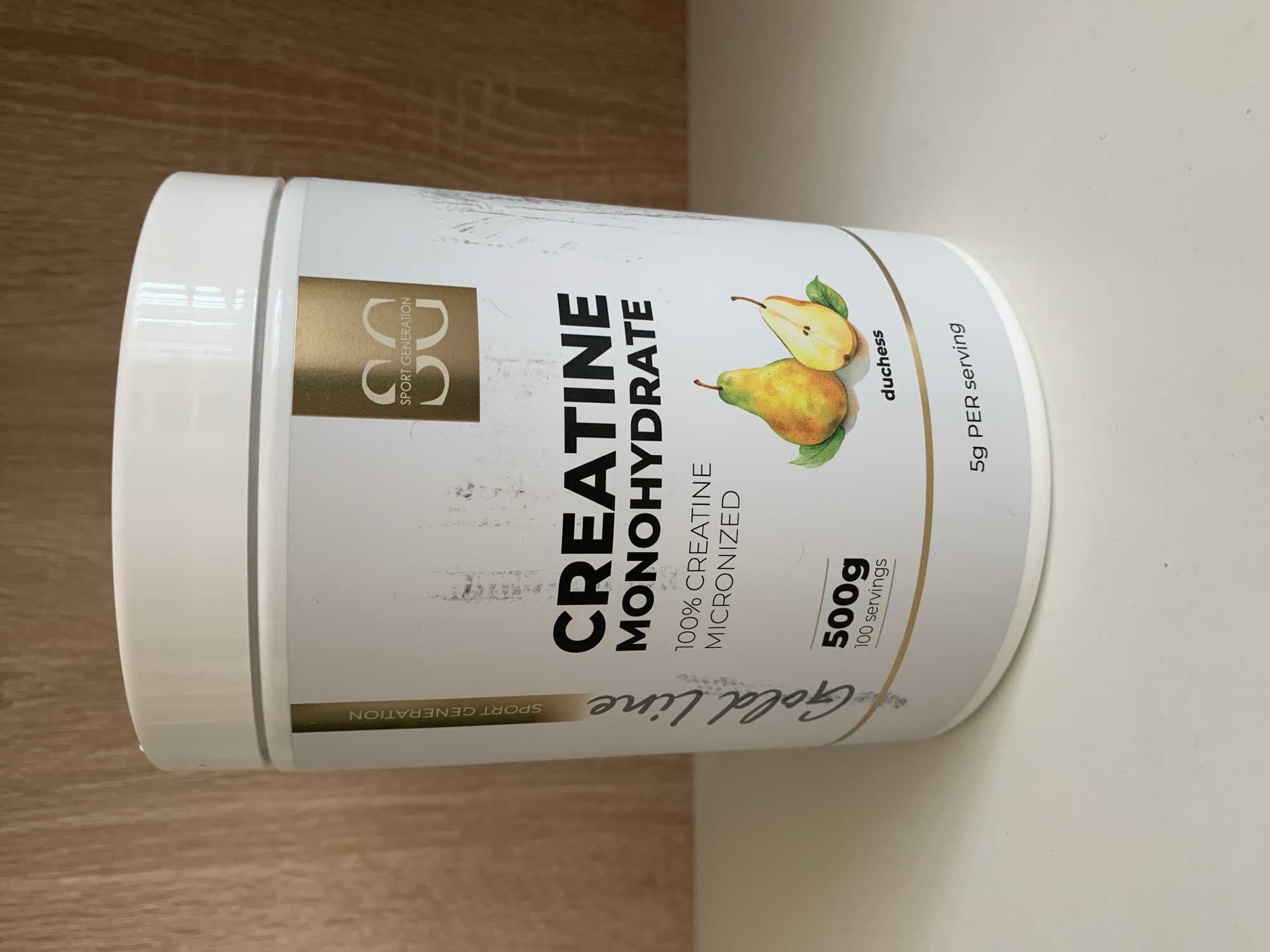 This is a close-up, eye-level, product shot of a white plastic tub of "Gold Line Creatine Monohydrate" supplement. The tub is positioned on a light-colored wooden surface, likely a table or shelf, against a plain white background. The lighting appears to be even and bright, suggesting an indoor setting, possibly during daylight hours.
The prominent text on the tub reads "CREATINE MONOHYDRATE" in large black sans-serif font, with smaller text below stating "100% CREATINE MICRONIZED". To the left of the main text, a gold square contains a stylized "SG" logo with "SPORT GENERATION" written vertically to the left of the letters. To the right of the main text, an illustration depicts two pears, one whole and one halved, with green leaves. Below the pears, the word "duchess" is printed vertically. Further to the right, "5g PER serving" is written vertically. Towards the bottom of the tub, "500g" and "100 servings" are displayed prominently, and a gold banner at the very bottom has "SPORT GENERATION" written horizontally. The tub itself is mostly white with gold and black accents, giving it a clean and professional appearance. There are some minor scuff marks on the surface of the tub, indicating it is a used product. The overall scene is still and focused solely on the product.
