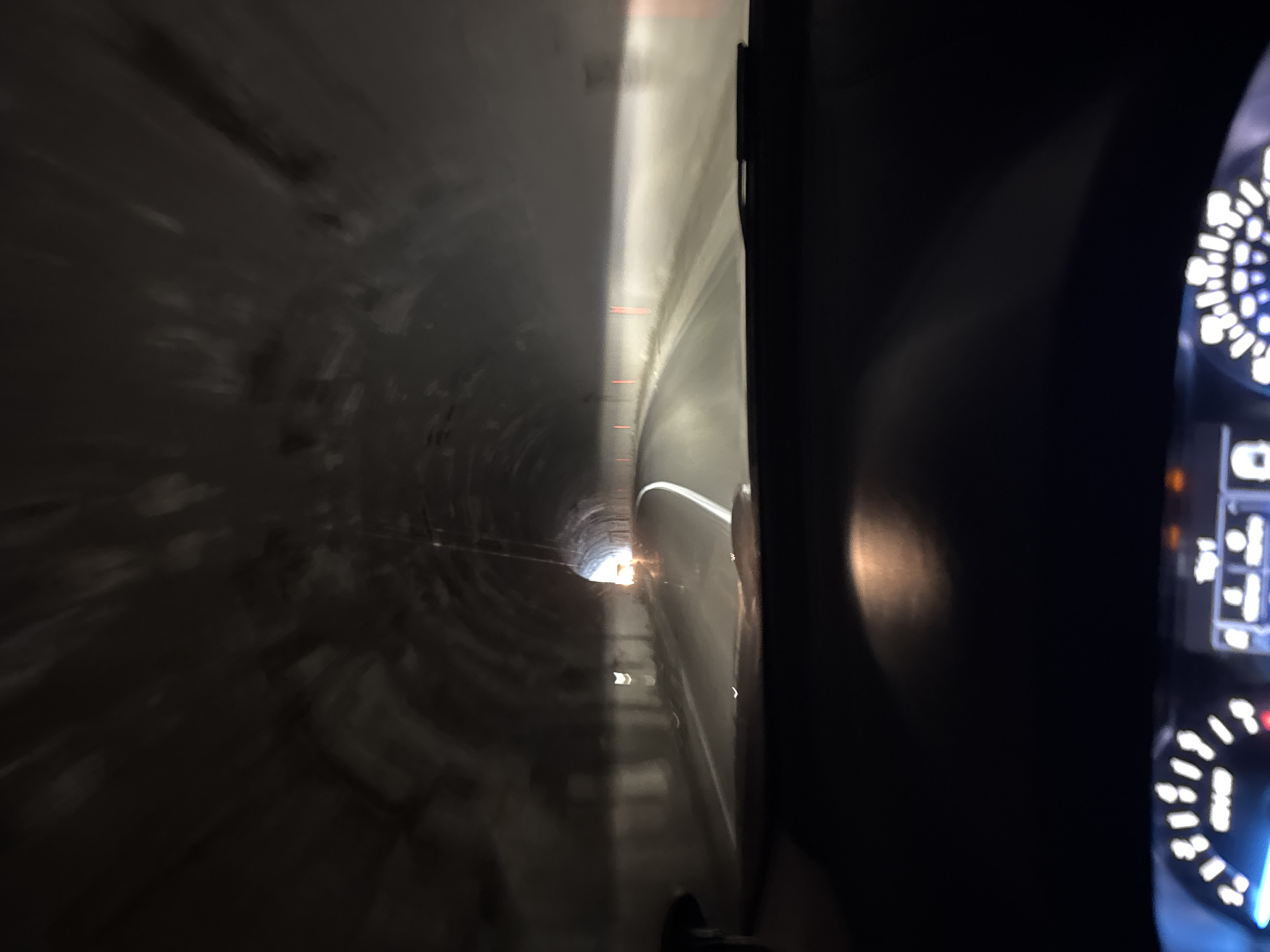 The image shows the interior of a vehicle moving through a tunnel, likely taken from the driver's perspective. The dashboard and instrument panel of the car are visible on the right side of the frame, illuminated by white and blue lights. The tunnel stretches out ahead, with light visible at the far end. The tunnel walls are concrete, and there are faint red lights along the upper portion of the wall. It cannot be determined that the tunnel is in Zagrađe, Montenegro.