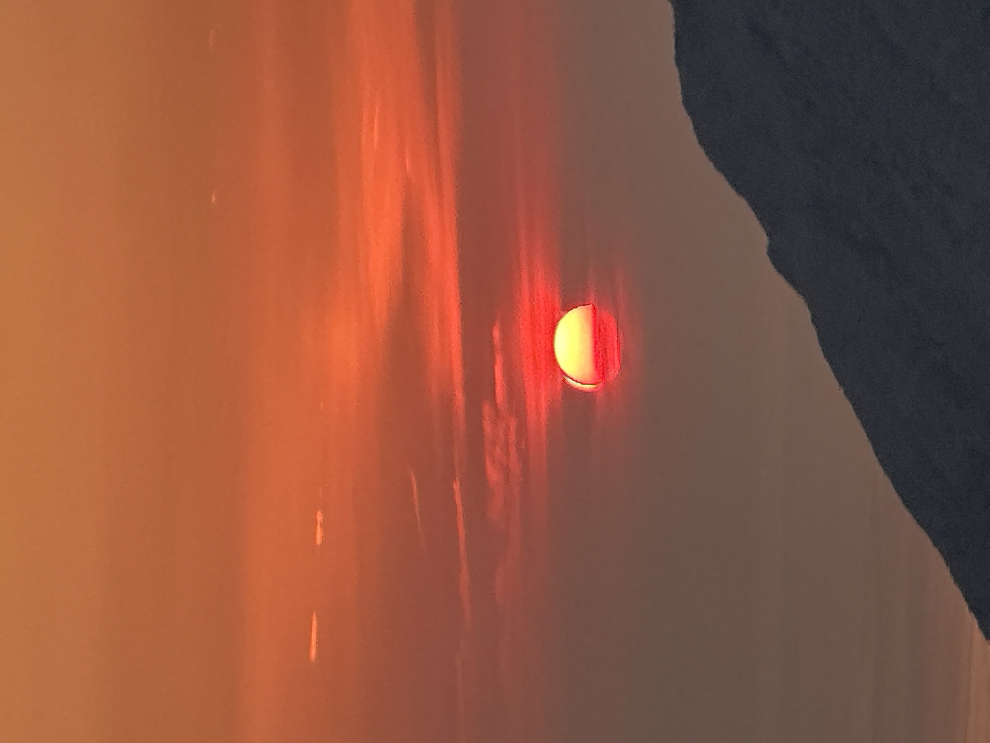 The image depicts a vibrant sunset over a mountainous landscape in Gluhi Do, Montenegro. The sky is a gradient of fiery reds and oranges, with wispy clouds streaked across the horizon. The sun, partially obscured by the atmosphere, appears as a bright, red-yellow orb on the left side of the frame, casting a warm glow. In the foreground, a dark, silhouetted mountain ridge cuts diagonally across the lower portion of the image, its textured surface suggesting rock and vegetation. The overall impression is one of natural beauty and tranquility, with the setting sun creating a dramatic and awe-inspiring scene.
