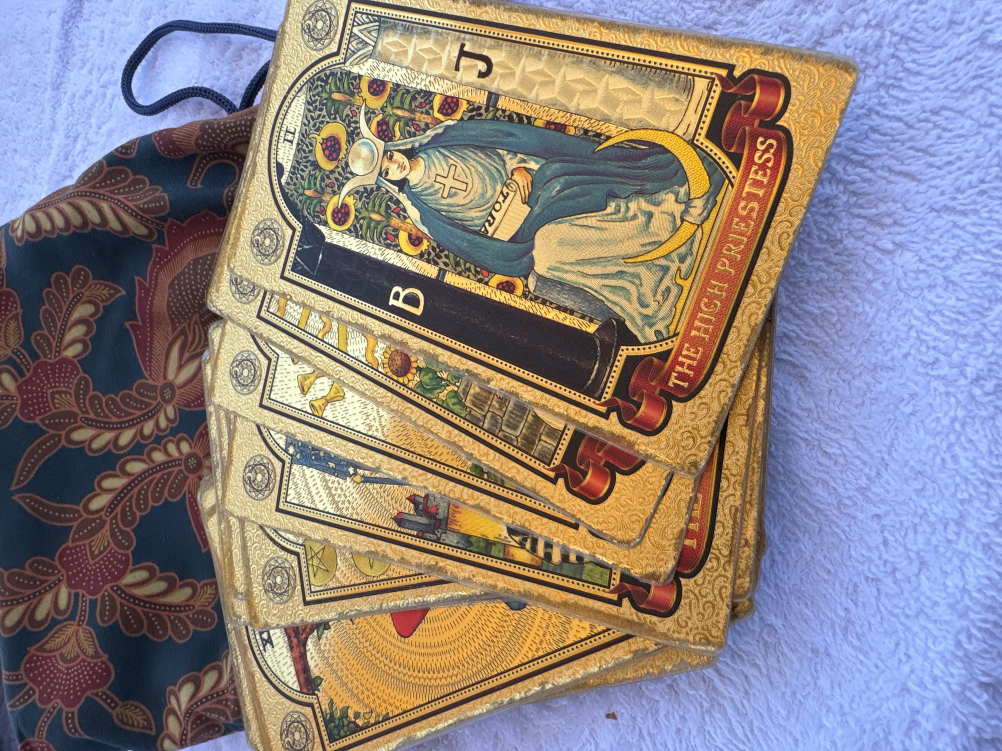 A deck of gold-edged tarot cards, topped by "The High Priestess," lies on a white, textured surface alongside a drawstring bag with a dark green and reddish-brown floral pattern. The cards are stacked at an angle, revealing glimpses of other cards in the deck. The scene is brightly lit. There are no people visible. The location is not specified.