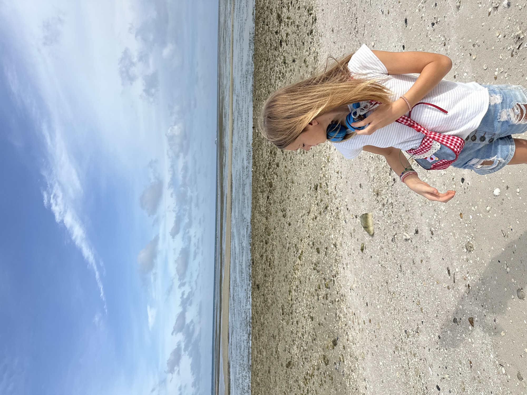 A young person with long, blonde hair stands on a sandy beach in ตำบลเกาะพะงัน, Thailand. They are wearing a white striped t-shirt, ripped denim shorts, and a small red and white checkered bag slung across their body. Blue headphones are held around their neck, and they wear bracelets on both wrists. The beach is covered in small shells and pebbles, with patches of shallow water and seaweed visible in the distance. The sky above is partly cloudy, with a calm sea stretching to the horizon.