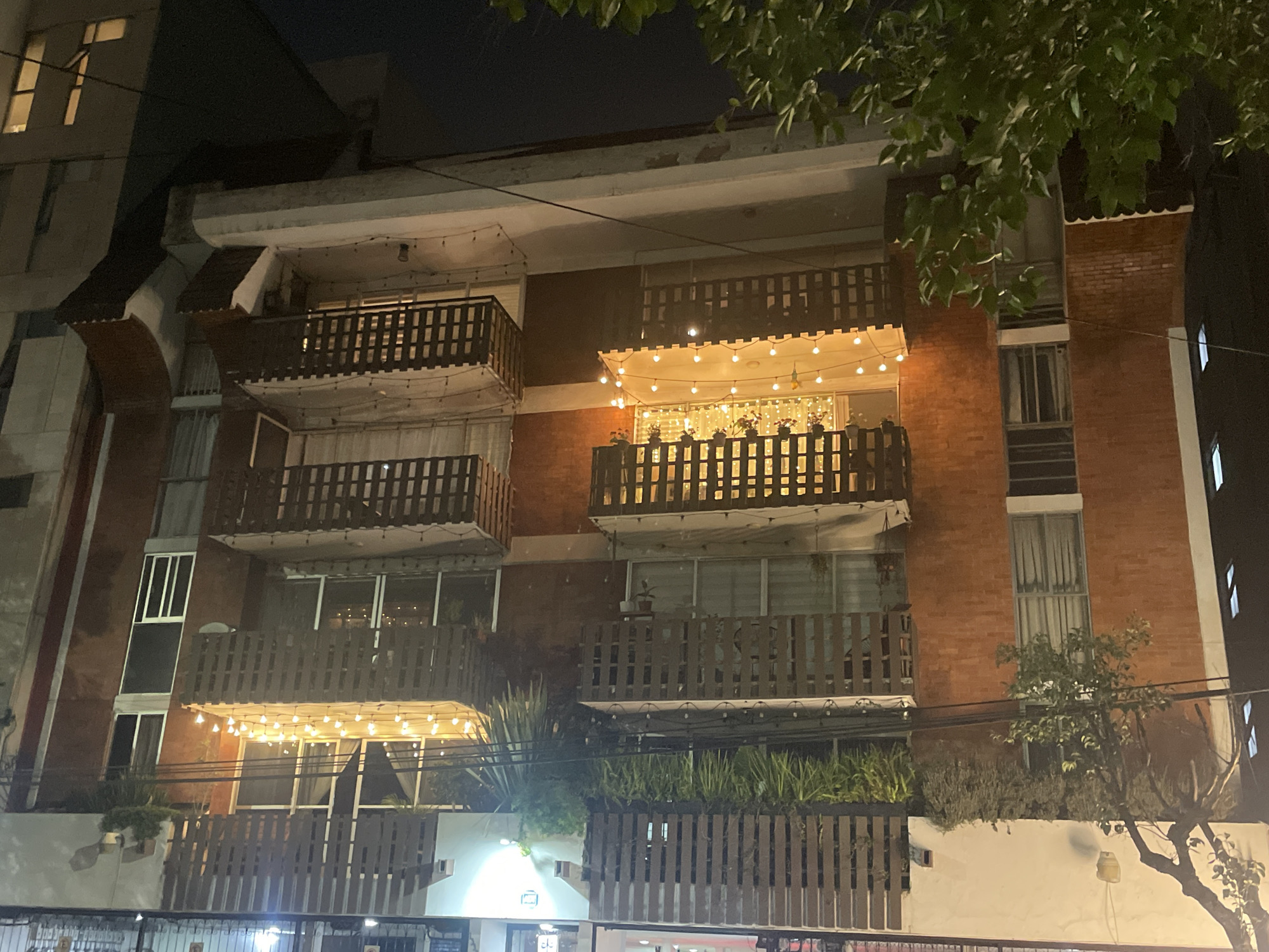 At night, a multi-story building in Mexico City, Mexico is visible. The building features red brick accents, interspersed with white sections. Each floor has a balcony with a dark brown wooden railing. String lights illuminate the balconies on the top and bottom floors. Plants and greenery are visible in the railing of the bottom balcony. The area is lit, with a tree obscuring the upper-right portion of the building. Another building is visible to the left, partially out of frame.