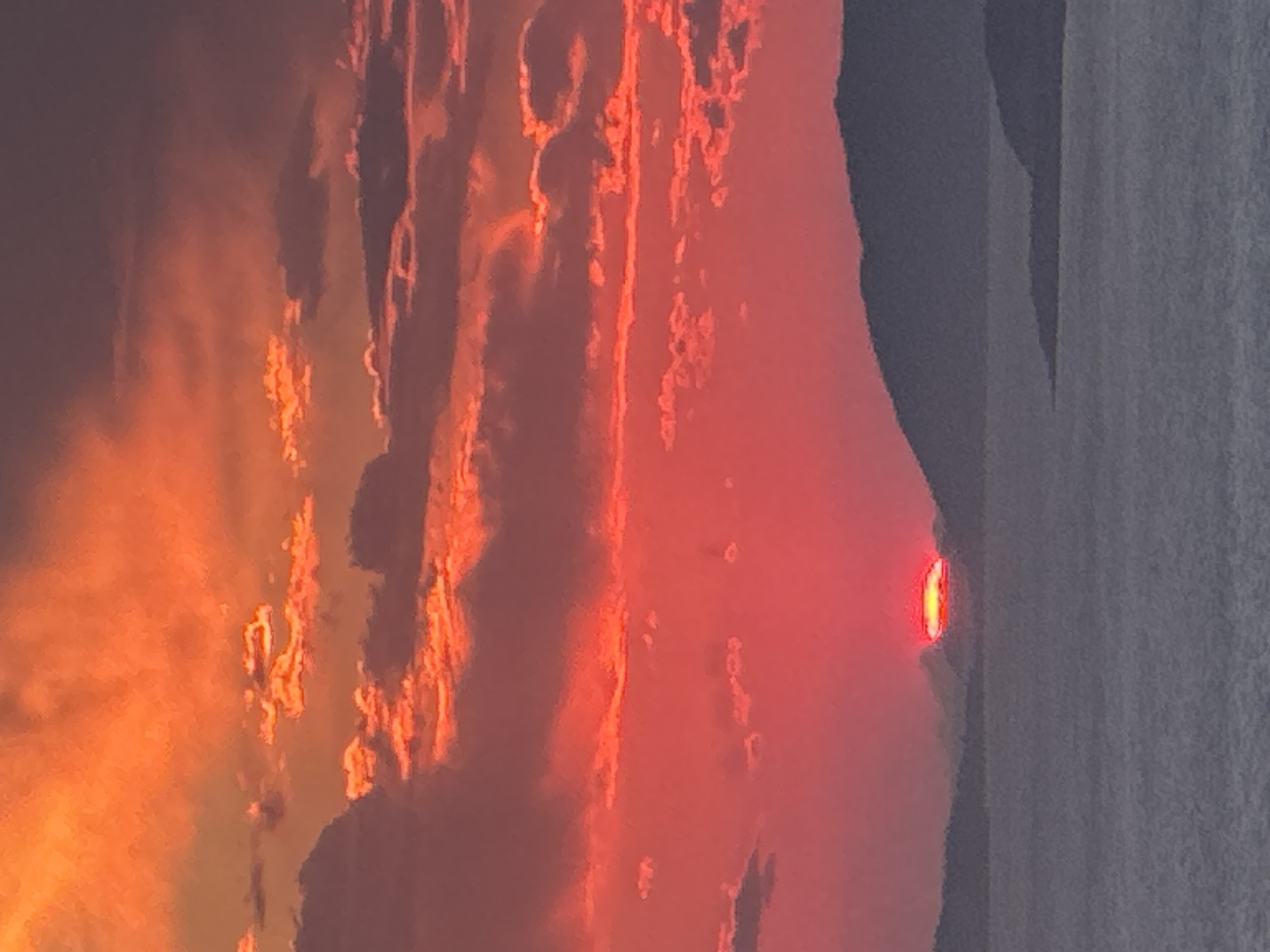 The image depicts a vibrant sunset over the Adriatic Sea, viewed from the coastal town of Zagora, Montenegro. The sky is a dramatic canvas of fiery oranges and deep reds, with scattered clouds tinged with the same intense hues. Silhouetted against this brilliant sky are the dark, undulating forms of distant mountains and islands. The sea below is a muted gray, reflecting the fading light and creating a stark contrast with the luminous sky. The sun itself is a bright, horizontal sliver peeking from behind a mountain ridge, casting a golden glow on the horizon. No people or man-made structures are visible, emphasizing the natural beauty of the scene.
