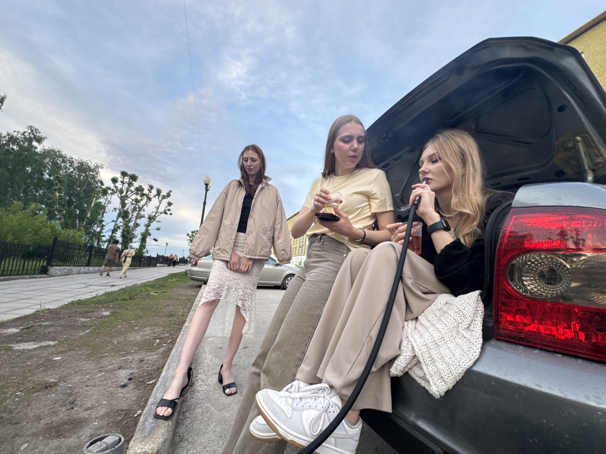 Three young women are gathered around the open trunk of a gray car. One woman is sitting in the trunk, smoking a hookah. Another woman is seated beside her, holding a drink in a clear plastic cup. The third woman is standing beside the car, leaning against the curb. The women are dressed casually. There are other people and cars in the background, and the city of Perm, Russia can be seen in the surrounding environment.