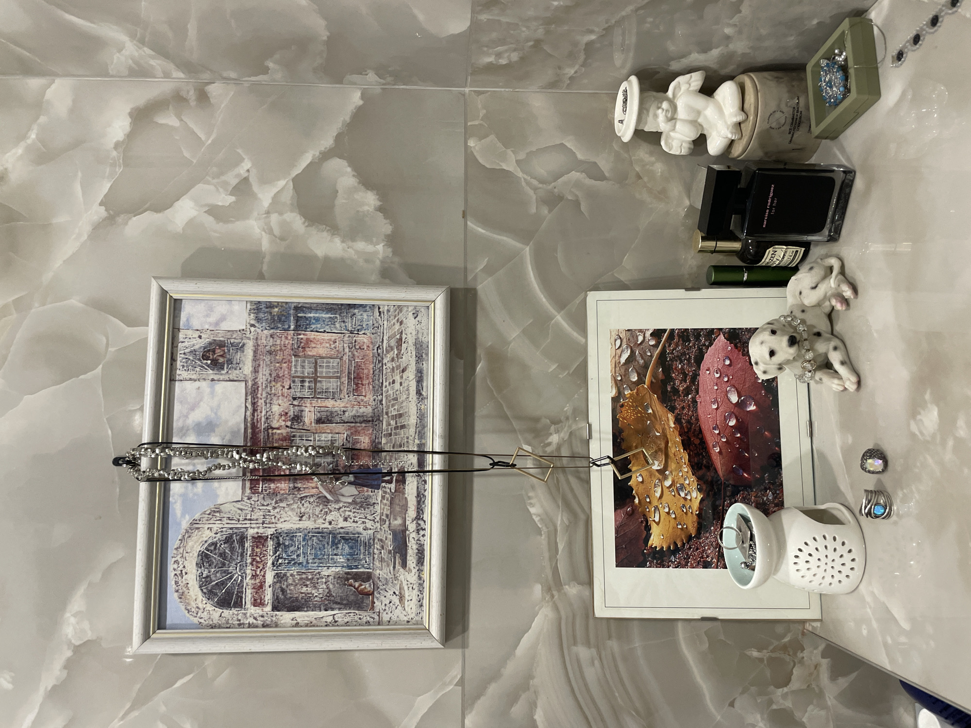The image shows a collection of items arranged on a surface with a marble-like pattern, likely a countertop or shelf. The objects include: two framed pictures (one depicting a building and the other showing leaves with water droplets), a necklace hung between the frames, a small white angel figurine, a gray candle holder, a box containing jewelry, several perfume bottles, a small Dalmatian figurine, a white ring holder, and a couple of rings. The items appear to be arranged for display or storage. Based on the user's prompt (city of Krasnoyarsk, Russia), this is potentially in a home in that city, but this cannot be definitively confirmed from the image alone.