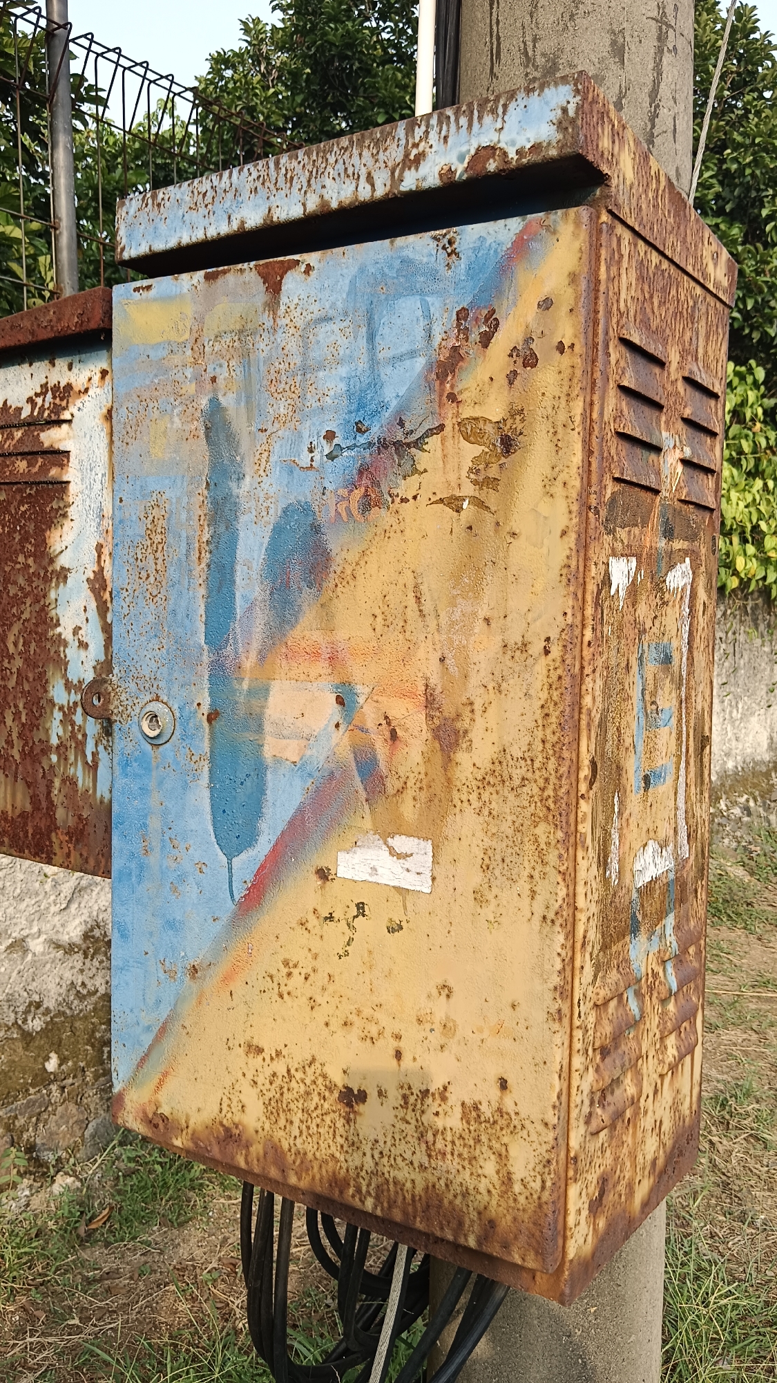 This is a close-up, eye-level, full-shot photograph of a rusted electrical cabinet mounted on a concrete utility pole. The cabinet is predominantly a weathered yellow color with significant rust patches and streaks. A portion of the cabinet door, painted blue, is open, revealing more rust underneath. There are remnants of spray paint graffiti in blue, red, and yellow on both the door and the main body of the cabinet. Some small, white rectangular stickers are also visible. Vents are present on the right side of the cabinet. Black electrical cables are bundled and emerge from the bottom of the cabinet. The cabinet is situated outdoors, with a blurred background of green foliage and a fence. The overall impression is one of decay and neglect. The location is identified as Kemang, Indonesia. There are no people or animals visible in the image.