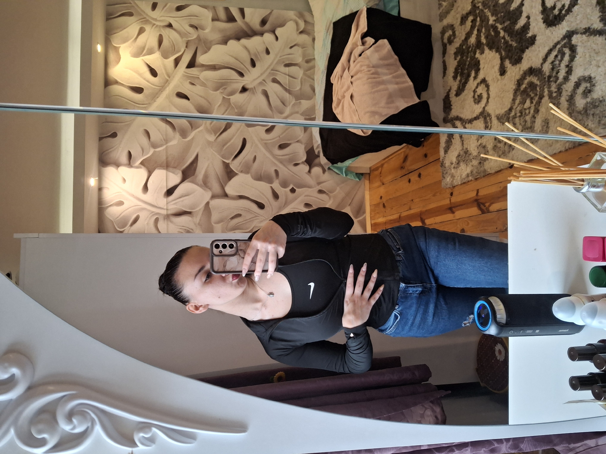 A young woman is taking a selfie in a mirror in a bedroom in Buhovo, Bulgaria. The woman is wearing a black Nike-branded top and blue jeans. The mirror reflects a wall with a decorative leaf pattern, a bed with pillows and blankets, a white dresser, and a speaker device. The environment includes wooden flooring, a rug, and various items on the dresser such as markers and a diffuser.