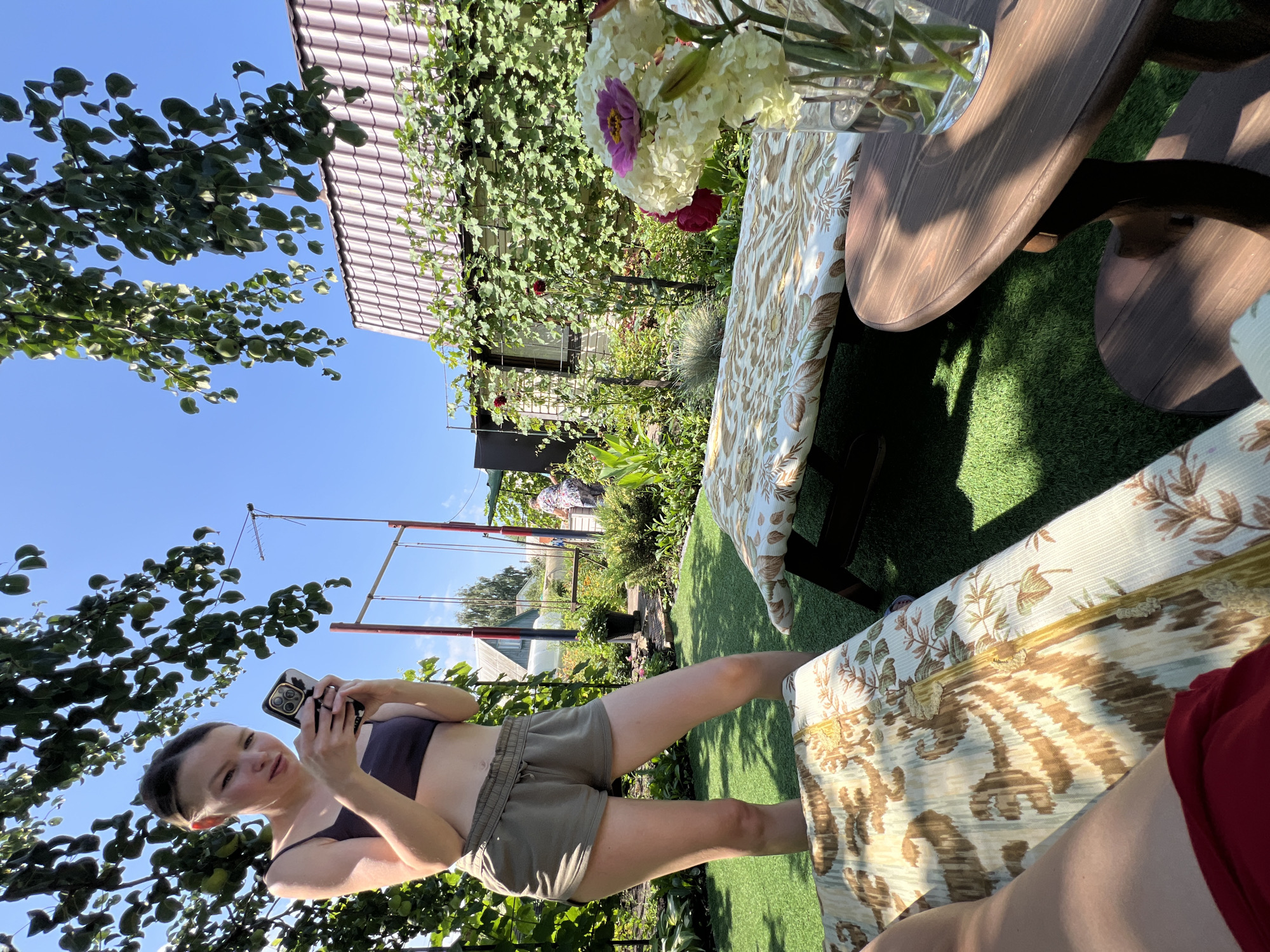In a backyard in Lopatino, Russia, on a sunny day, a young woman stands holding a smartphone, seemingly engaged with its screen. She wears a dark purple crop top and olive green shorts. To her left, a patterned lounge chair is partially visible. In the background, a swing set stands near a building with a red-tiled roof, surrounded by lush greenery and flowers. Another person is visible in the distance near the swing set, and a vase of white flowers sits on a wooden table in the foreground. The ground is covered with artificial turf, and the scene is shaded by overhanging tree branches.