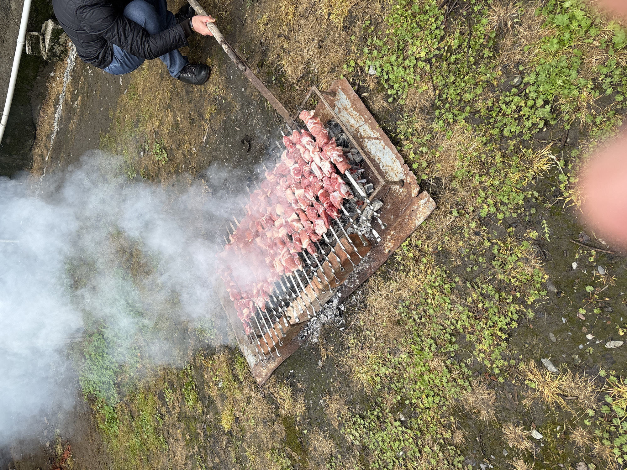 The image shows an outdoor barbecue scene, possibly in a backyard, in Kartubani, Georgia. Skewers of raw meat are arranged on a charcoal grill. Smoke rises from the grill, obscuring some of the ground. A person wearing a dark jacket and jeans sits nearby, holding a long metal rod. The surrounding area is covered in grass and patches of bare earth. Another hand and finger are partially visible in the foreground. A building is to the left.