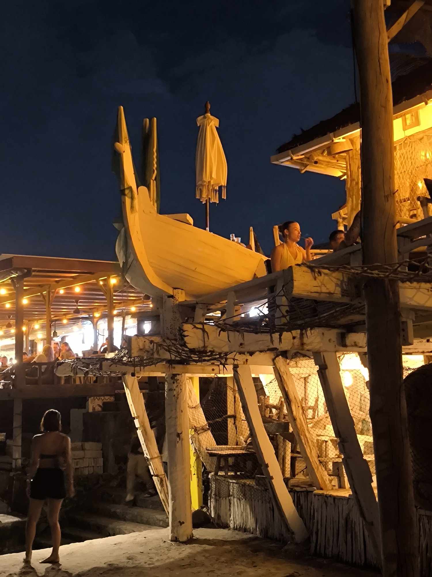 At night in Canggu, Indonesia, a woman stands in the foreground on a sandy surface. She is facing away from the camera, wearing a dark, two-piece outfit. Behind her are stairs leading to a partially visible wooden structure that supports a decorative boat. The boat appears to be part of a bar or restaurant, as people are seated on the elevated structure. Lighting is provided by strings of lights, illuminating the scene. The structure is made of wooden beams and appears to be built over the beach.