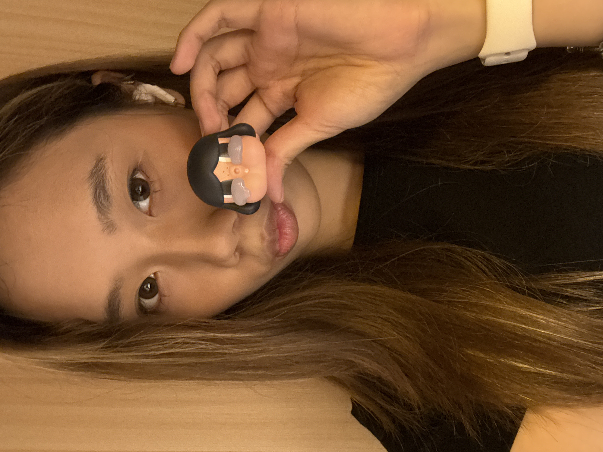 A person with long brown hair is lying on their side on a light brown surface. They are wearing a black top and a white smartwatch. They are holding a small, stylized figure in their hand, positioning it near their lips. The figure has black hair and a peach-colored face. A hair clip is visible in the person's hair. The location cannot be determined.