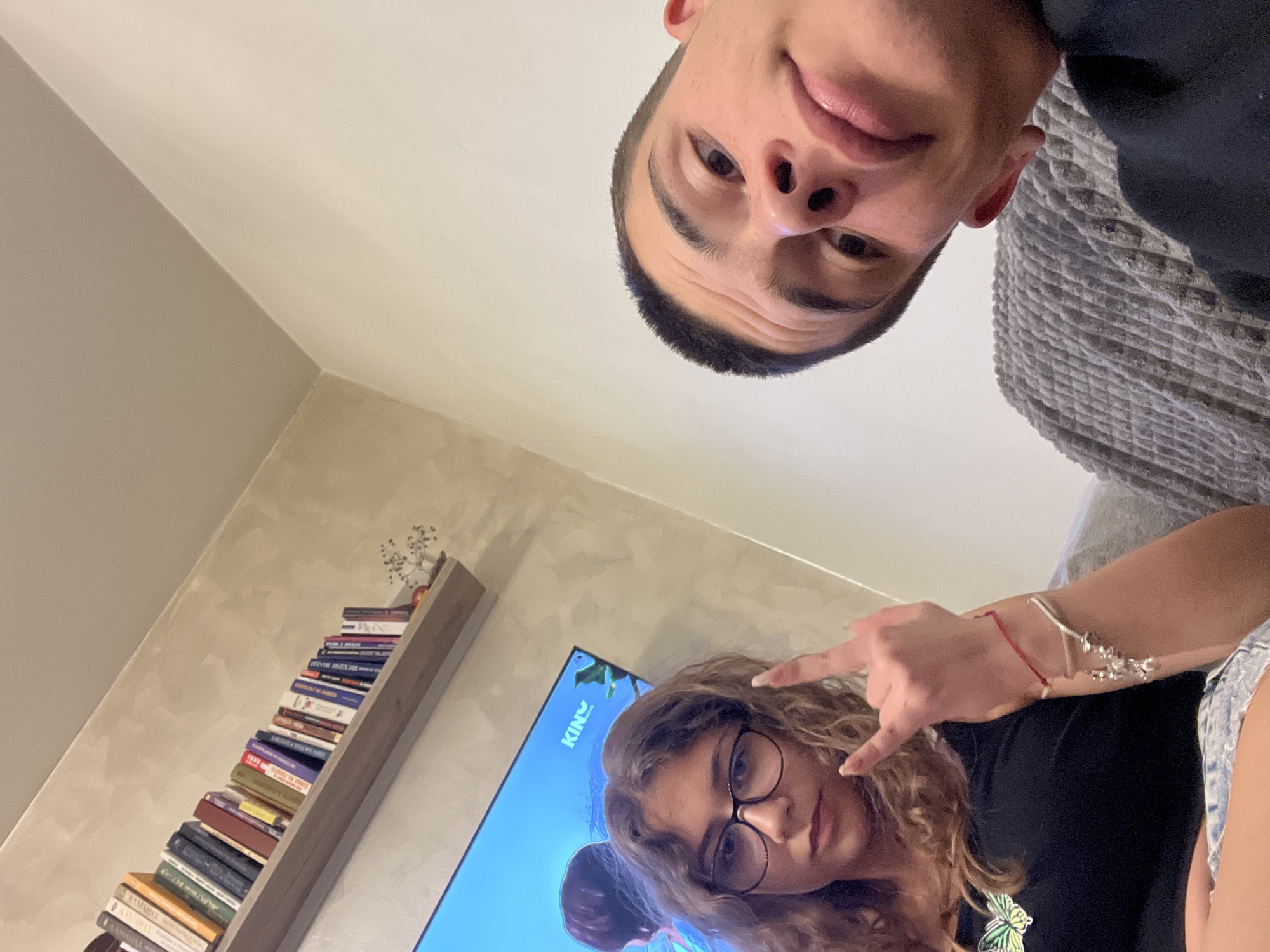 Two people are in a room. One, a fair-skinned male, is positioned above the other, facing the camera, wearing a dark shirt, and is near a gray blanket. The other, a fair-skinned female with curly hair and glasses, is below, facing the camera, wearing a black shirt, blue denim shorts, and making a rock-and-roll hand gesture. A bookshelf with books and a TV screen showing animation are visible in the background. The location cannot be identified as Plovdiv, Bulgaria, based on the visual information provided.