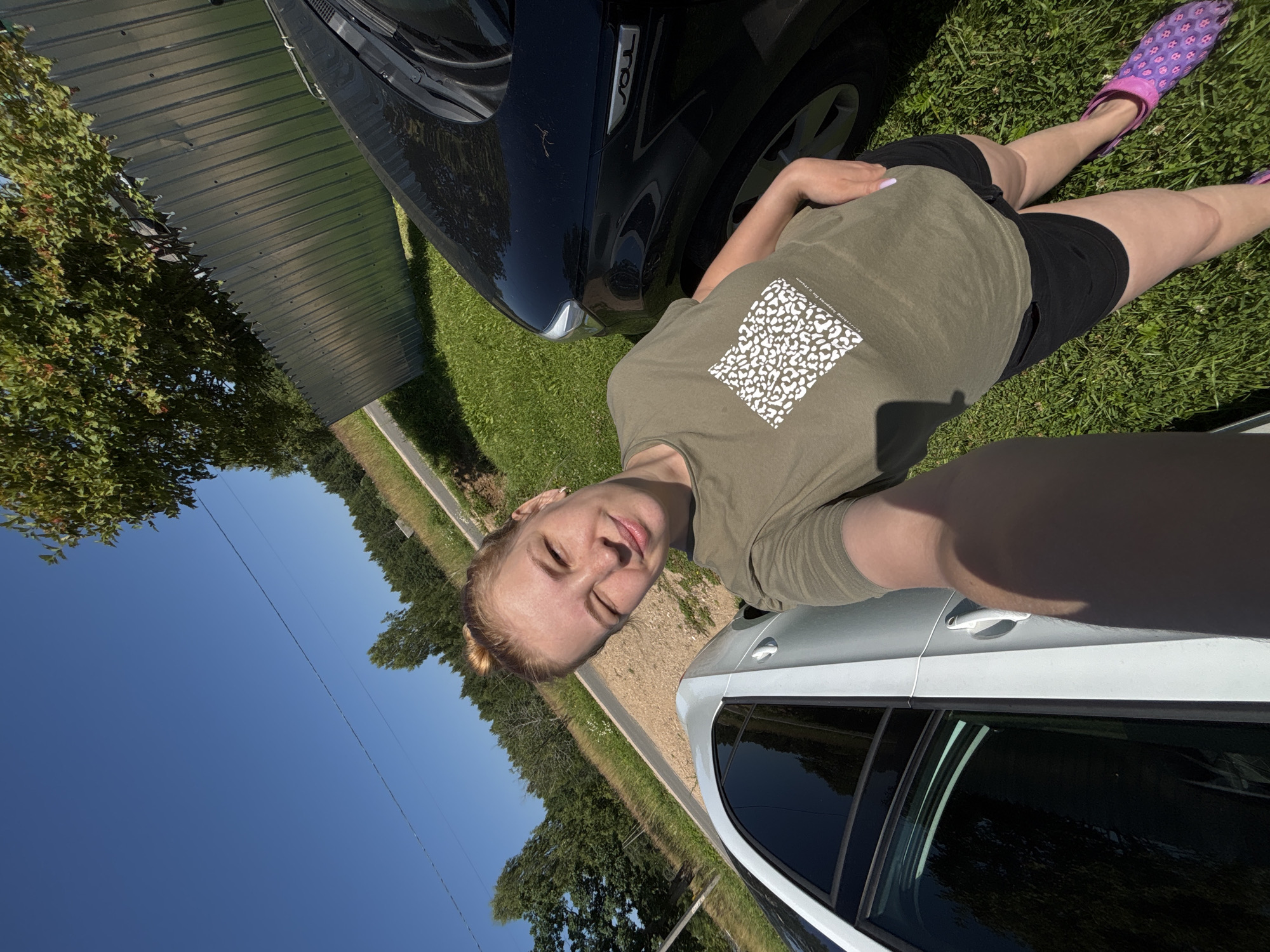 A woman is taking a selfie outdoors. She is sitting on a grassy area next to a black car and a silver car. She is wearing a green t-shirt with a white pattern and black shorts. Her right foot, wearing a purple clogs, is visible. The scene appears to be set during daytime with clear blue skies. In the background, there is a metal fence and trees. The woman has light brown hair pulled back and is smiling at the camera, showing a relaxed and happy emotion. The word "SOUL" is visible on the black car. The location is identified as Dunilovo, Russia.