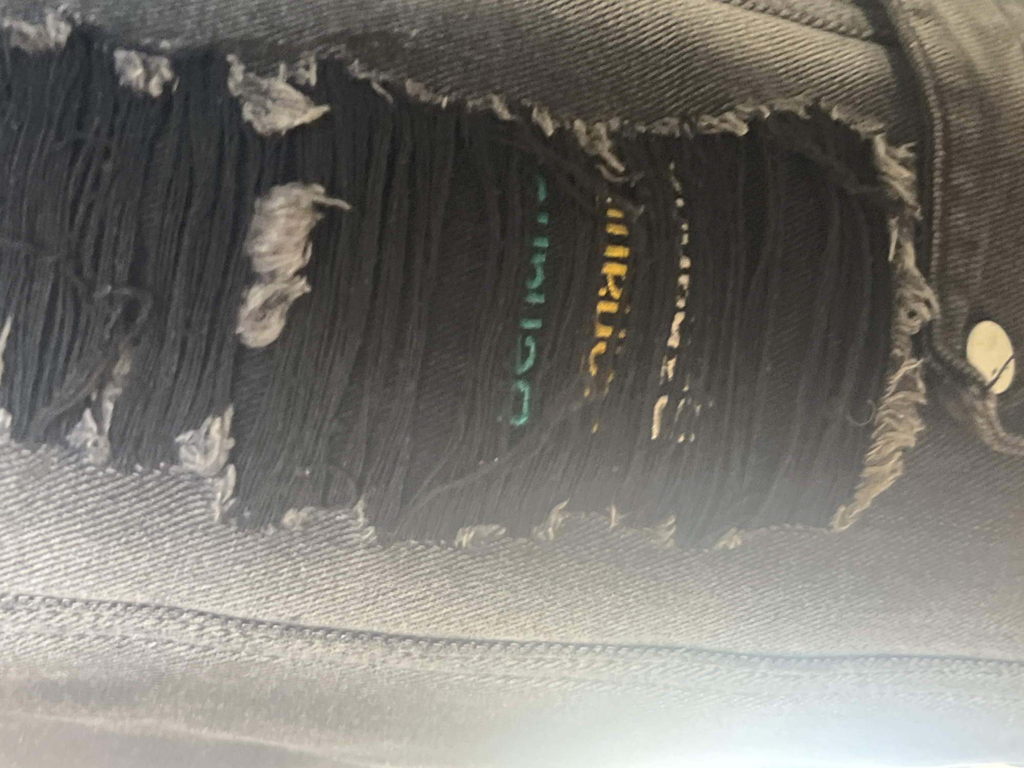 The image shows a close-up view of ripped denim jeans. The focus is on a distressed area of the fabric, where threads are frayed and hanging loosely, revealing a black material underneath. On this black material, there is visible text printed in green and yellow. The green text appears to read "SUR", while the yellow text is harder to decipher but seems to contain letters like "RU" and "C". The denim itself is a dark grey or black color, suggesting a weathered or intentionally distressed style. The lighting is somewhat diffused, and there are no clear indications of time of day or weather. The background is out of focus, but it appears to be the continuation of the same denim fabric. The image is likely a detail shot of clothing, possibly for a fashion advertisement or a personal item. No people are visible in the frame.