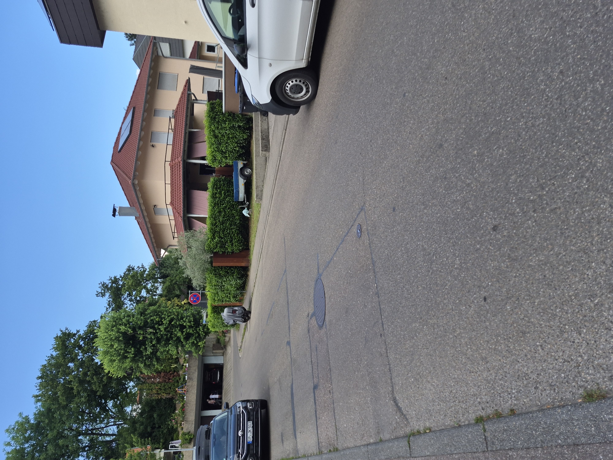 In a residential area, a black Volkswagen T-Roc with license plate "GP JK 503" is parked on the left side of a paved road. A white van is parked on the right side of the road. A covered scooter is parked on the sidewalk. A small blue trailer is visible behind a hedge. In the background, a beige house with a red tiled roof and solar panels is visible, along with lush green trees and a clear blue sky. The road shows visible lane markings and a manhole cover.