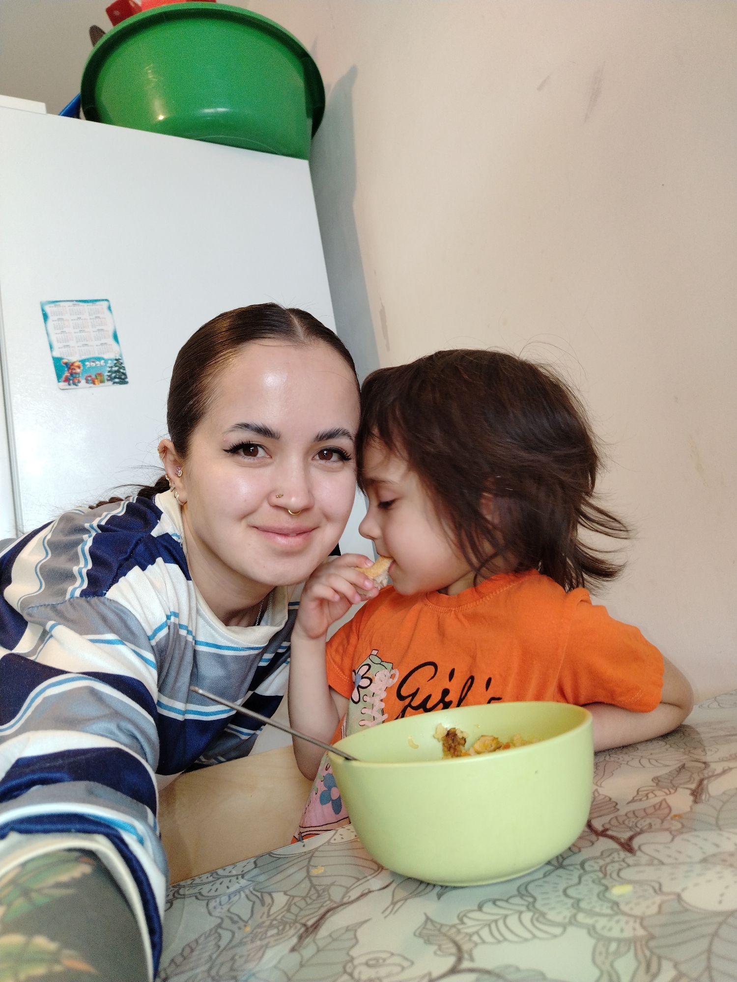 Captured indoors, the photograph depicts a fair-skinned adult female with dark hair pulled back, wearing a horizontally striped blue and white top. She smiles at the camera. Adjacent to her is a young child with dark, shoulder-length hair, clad in an orange t-shirt. The child is in the act of eating something. Between them sits a light green bowl containing food. The table is covered with a patterned tablecloth. In the background, a white refrigerator is visible with a green plastic bowl atop it. A calendar is attached to the refrigerator. The location is Noviy, Russia.