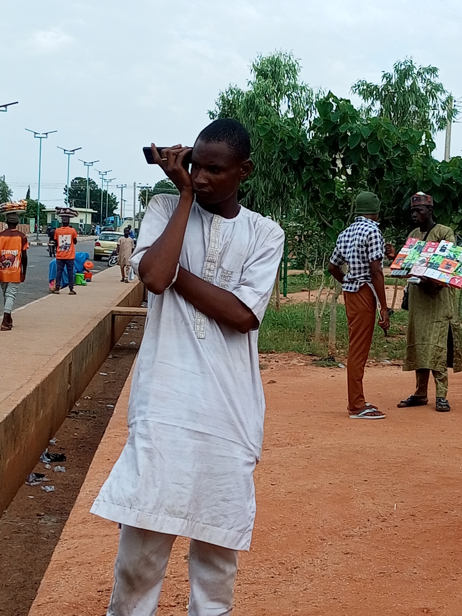 In an outdoor urban setting, a young man in a white garment is holding a black mobile phone to his ear, seemingly engaged in a conversation. He stands on a reddish-brown dirt path, his posture suggesting casual engagement with the device. In the background, several other individuals are present. To the left, two men in orange shirts walk away from the camera, each carrying items on their heads. Further behind them, a man in a light brown robe walks alongside a yellow car. To the right of the main subject, two men are interacting: one in a checkered shirt and brown pants, and another in a long green garment, holding a colorful box of items that appears to be for sale. The environment is characterized by a paved walkway or road, a trench running alongside the dirt path, and lush green trees and vegetation in the background. The sky is overcast.