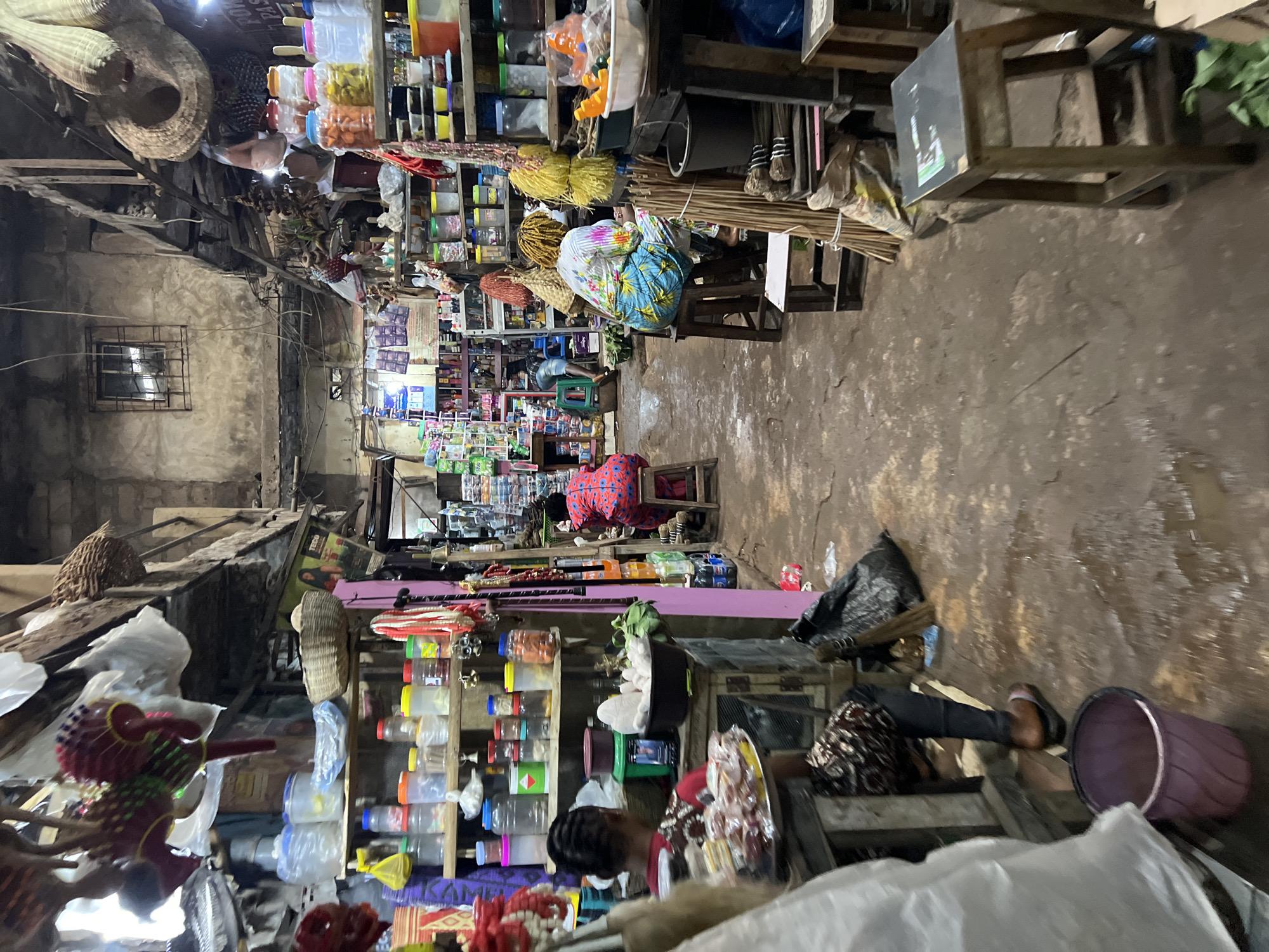 This image depicts an indoor market scene in Nkpor, Nigeria. The market appears to be a covered alleyway lined with stalls. The floor is made of packed dirt and appears wet in places. The stalls are filled with various goods, including plastic containers of food items, woven baskets, strings of beads, and other assorted merchandise.
In the foreground, a person wearing a patterned shirt and dark pants is seated on a stool, facing away from the camera. They are positioned behind a wooden counter displaying food items on a tray. To the left, a large purple bucket is visible on the ground.
Further into the market, another person is seated on a stool, wearing a red and blue patterned dress. They are also facing away from the camera. To their right, a person in a colorful floral blouse and blue patterned pants is seated on a stool, facing away from the camera.
The stalls are constructed from wood and metal, with shelves filled with goods. The ceiling appears to be made of corrugated metal and wood beams. The lighting is dim and appears to be a mixture of natural light from openings and possibly artificial lighting. The overall impression is of a bustling, open-air market.