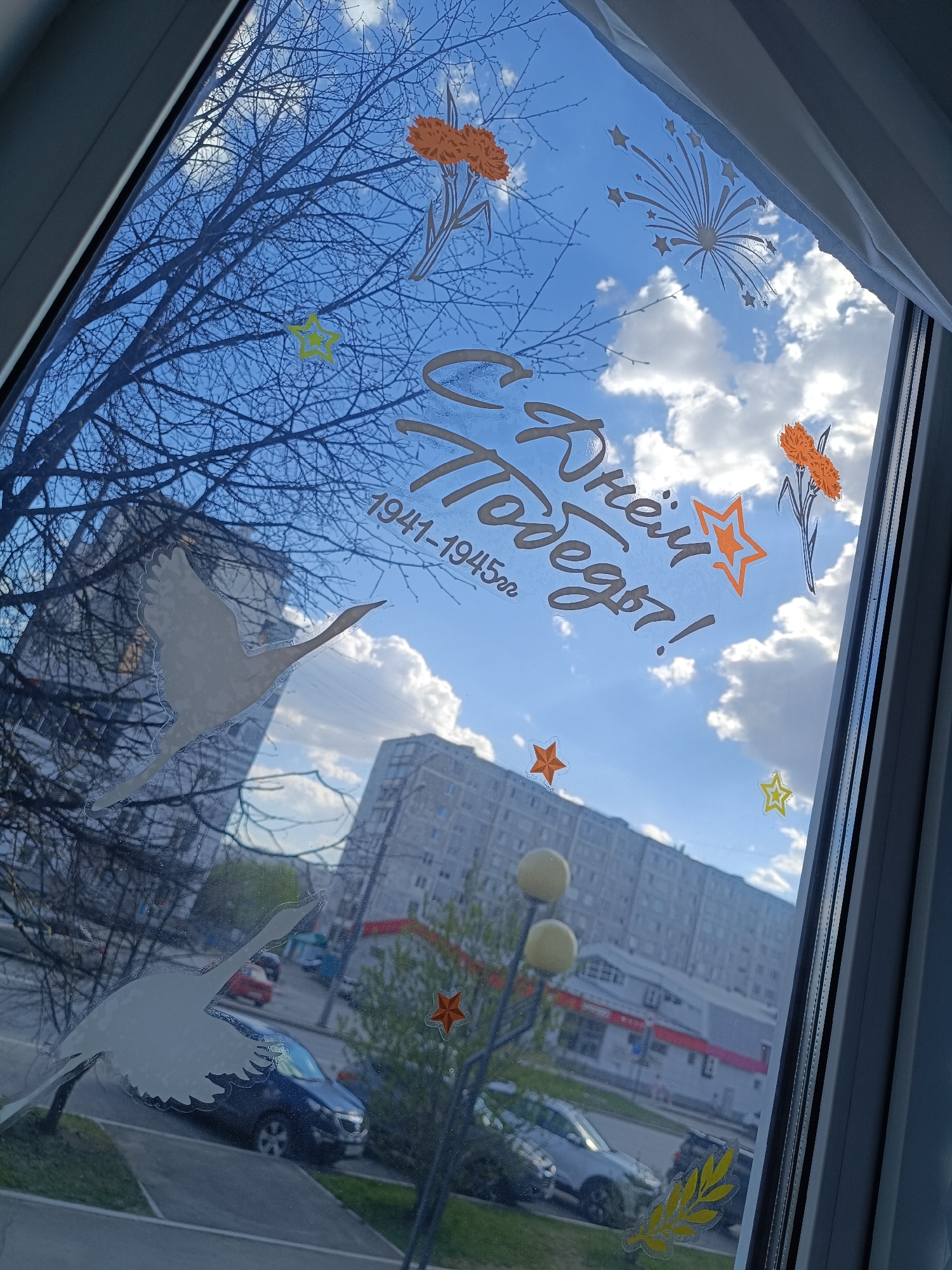 View through a window decorated with stickers commemorating Victory Day. The stickers include: two white swans, stylized text in Russian, orange flowers, orange and yellow stars, and a fireworks design. Outside the window is an urban scene: apartment buildings, trees with sparse leaves, cars parked on the street, a street lamp with globe-shaped lights, and a sidewalk. The sky is blue with fluffy white clouds. The location is likely in Tobolsk, Russia, given the Victory Day theme.