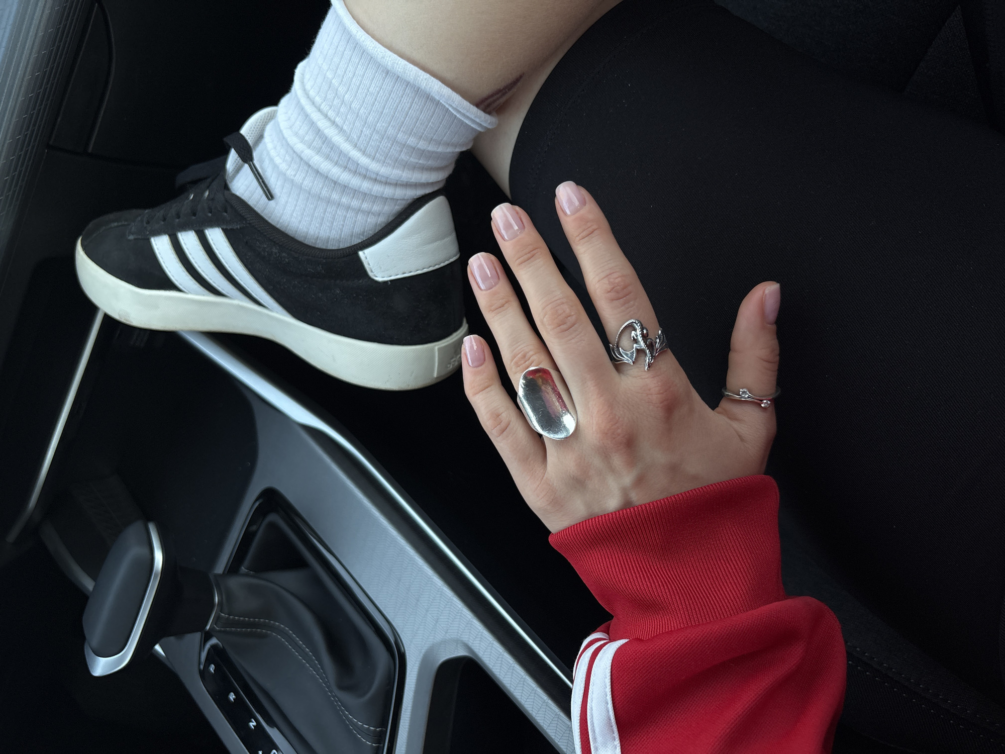 The image shows the interior of a car. A person's leg, clad in a white sock and black Adidas shoe, is resting on the center console. The person's hand is visible, adorned with three rings: a large, oval silver ring on the index finger, a dragon-shaped ring on the middle finger, and a thin ring with two small stones on the pinky finger. The person is wearing a red jacket with white stripes on the sleeves. The car's gear shift is visible in the lower left corner. The location is indeterminable from the image.