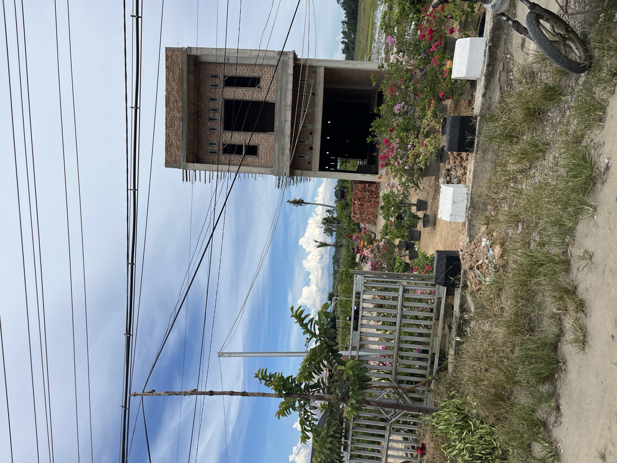 In Seuneubokpunti, Indonesia, a construction site features an unfinished two-story building made of brick and concrete. The building has multiple open windows and doorways, with scaffolding on one side. In front of the building, a metal fence encloses an area with potted flowering plants, including bougainvillea in vibrant pinks and reds. A young tree stands near the fence. Beyond the plants, a pile of bricks sits to the left, and a field with water is visible in the distance. Overhead, electrical wires crisscross against a cloudy blue sky. The foreground shows a dirt path with dry grass and a portion of a motorcycle on the right.