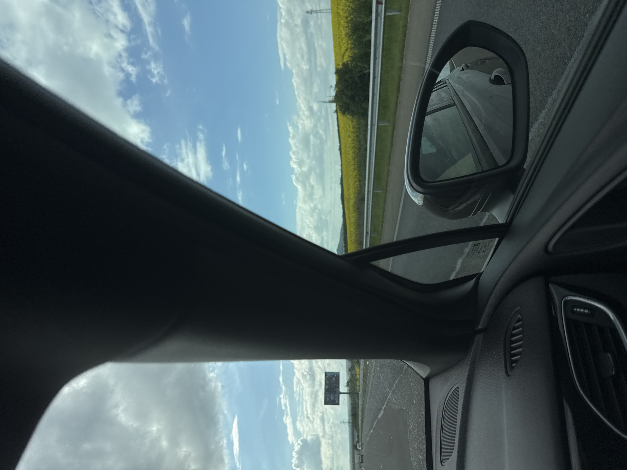 The view from inside a car, likely a passenger seat, traveling on a highway outside of what could be Bulgarovo, Bulgaria. The vehicle is moving past a field of yellow crops, visible through the side window and reflected in the side mirror. The sky is partly cloudy. Roadside barriers and a distant powerline are also visible. The car's interior is dark gray, with elements of the dashboard and the side mirror housing filling the frame. The photograph is taken at a tilted angle.