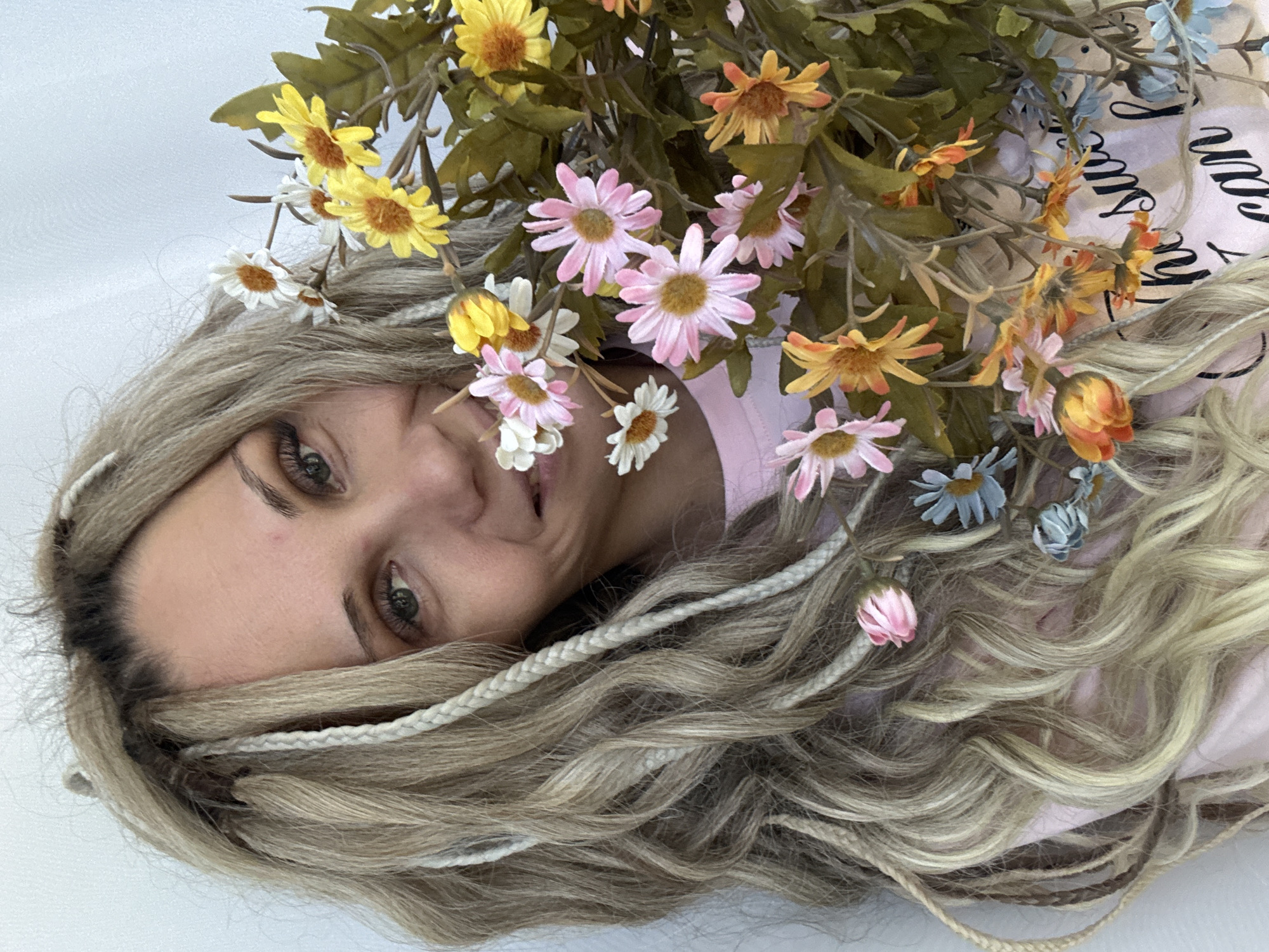 A woman with blonde hair and braids, partially covered in a bouquet of yellow, pink, orange, and blue flowers, is lying on a white surface. She is wearing a pink top. The location is likely a studio setting, given the neutral background. Rusanovo, Russia, is not visually confirmed.