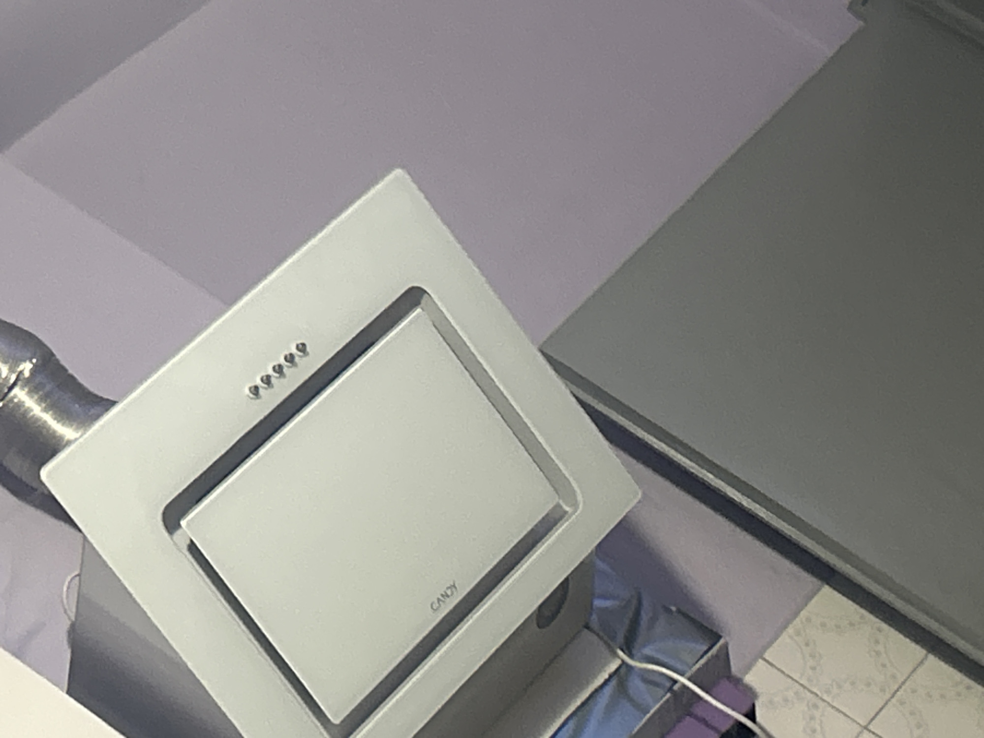 A white Candy brand range hood is mounted on a light purple wall. The range hood has a glass panel with several buttons on it. A silver metal duct is attached to the top of the range hood. To the left and below the range hood, a portion of a gray refrigerator is visible. Further down and to the left, some items in blue and purple are stacked, with a white cord extending from them. The flooring below appears to be tiled with a white and patterned design. The overall scene suggests a kitchen or dining area.