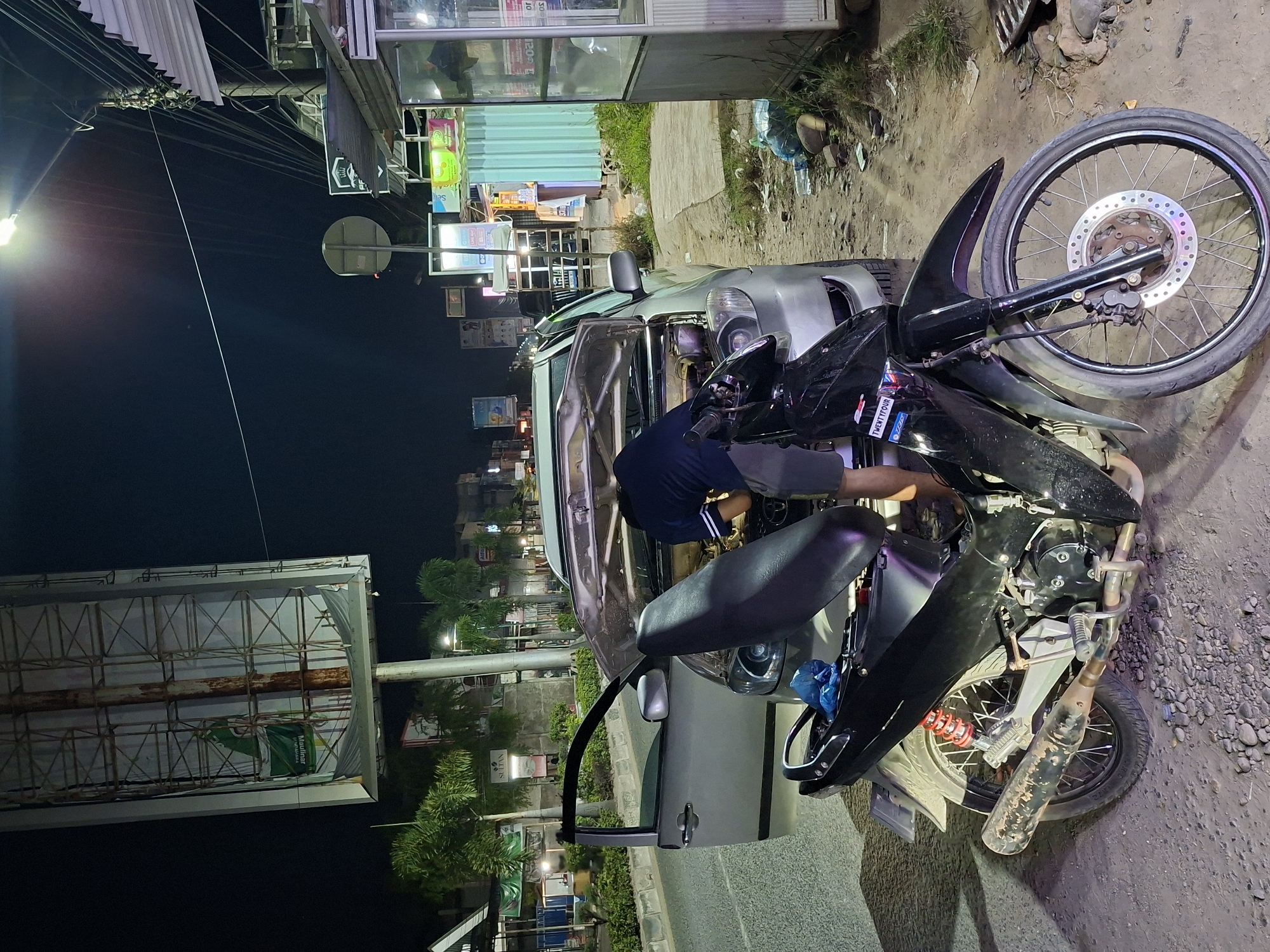 A nighttime scene shows a silver car with its driver's side door open. A man in a dark blue shirt is leaning into the car's engine compartment. Parked next to the car is a black motorcycle with a damaged front fender. The background includes urban elements like streetlights, storefronts with illuminated signs, roadside vegetation, and a large billboard structure. The setting is likely a city street in Lhokseumawe, Indonesia, given the information provided. The ground appears to be a mix of asphalt and gravel.