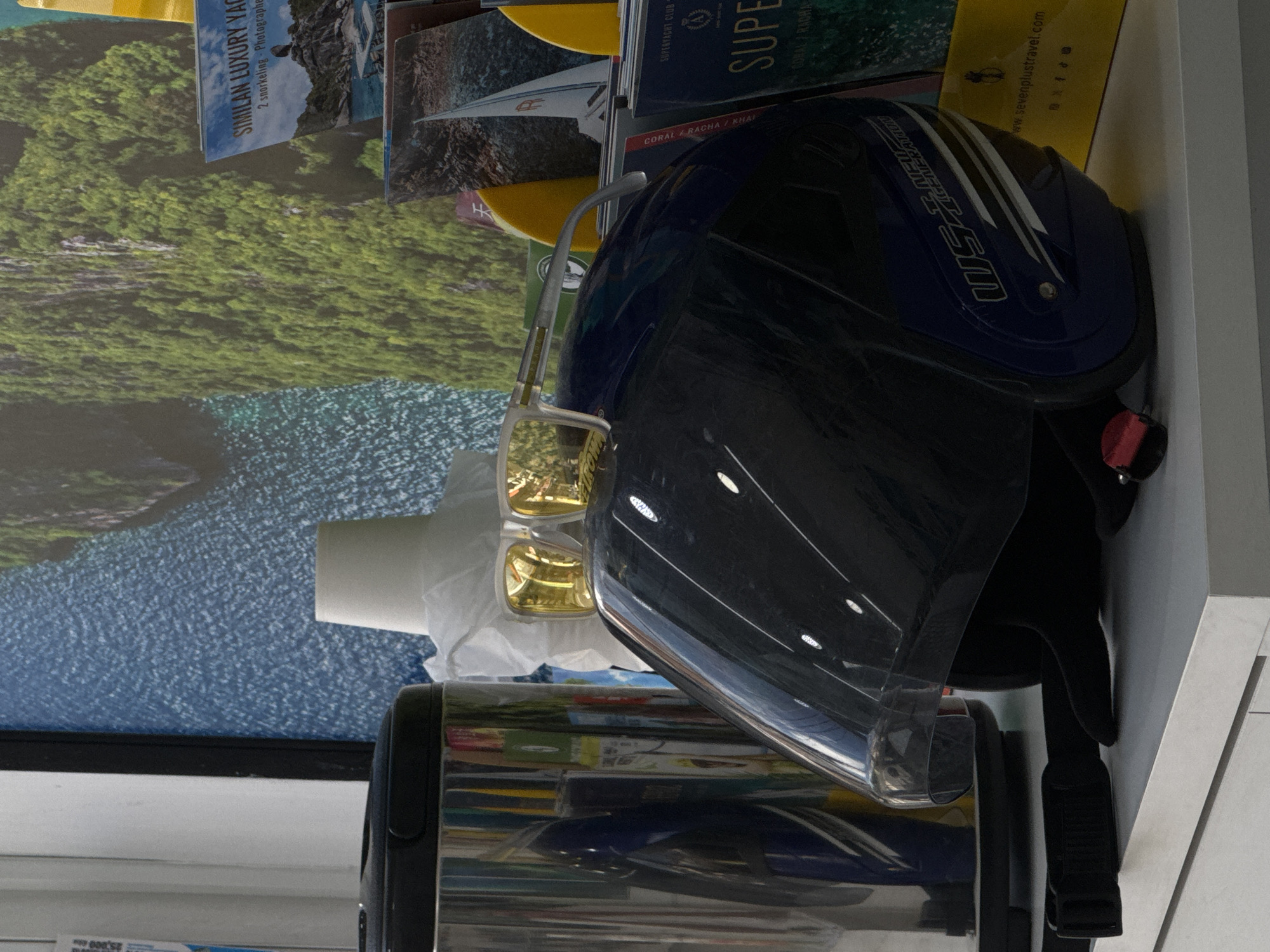 A motorcycle helmet with yellow tinted sunglasses is displayed on a white shelf in front of a wall covered with a mural of a tropical island. To the left of the helmet is a silver thermos. Behind the helmet, partially obscured, is a white paper cup. To the right of the helmet are several brochures and a yellow shopping bag with text that reads "www.sevenoplusltravel.com". The scene appears to be an indoor display, possibly in a shop located in Karon, Thailand.