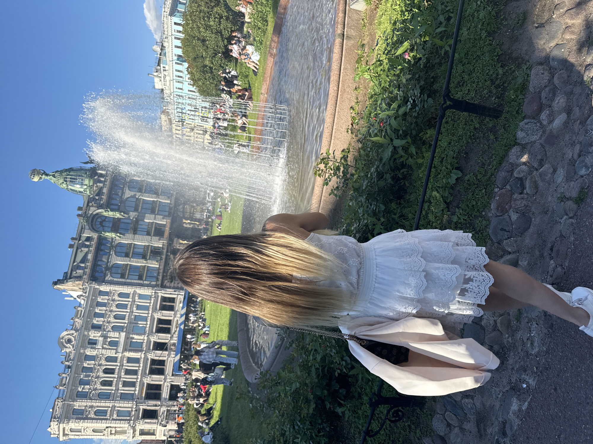 A woman is captured from behind, her long blonde hair cascading down her back as she stands near a fountain in a park. She is wearing a white lace dress and white sneakers, with a light pink jacket draped over her shoulder. The fountain is active, sending water spraying into the air. In the background, a large, ornate building with a green dome stands prominently. Several people are scattered across the green lawn, some sitting and others standing, suggesting a leisurely daytime activity. The sky is a clear, bright blue, indicating good weather. The scene appears to be Saint Petersburg, Russia, given the architectural style of the buildings. The overall mood is serene and pleasant, capturing a moment of enjoyment on a sunny day.