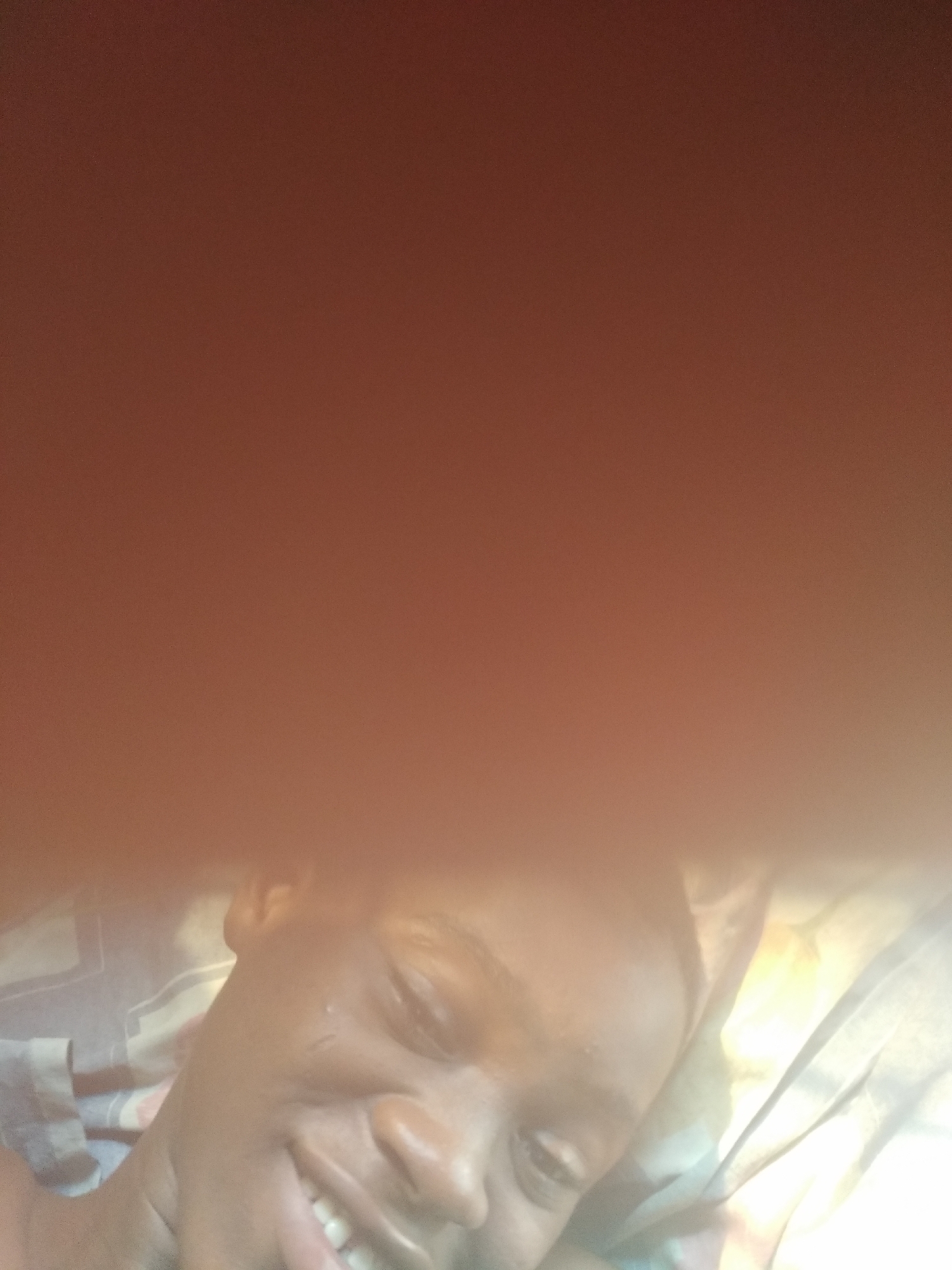 A close-up, poorly-lit photograph shows a person lying down, seemingly in a bed. The top portion of the image is obscured by an out-of-focus, reddish-brown area. The person's face is partially visible, showing a smile. The person appears to be lying on patterned bedding next to a white curtain. The location of Shangisha, Nigeria cannot be determined from the provided image.