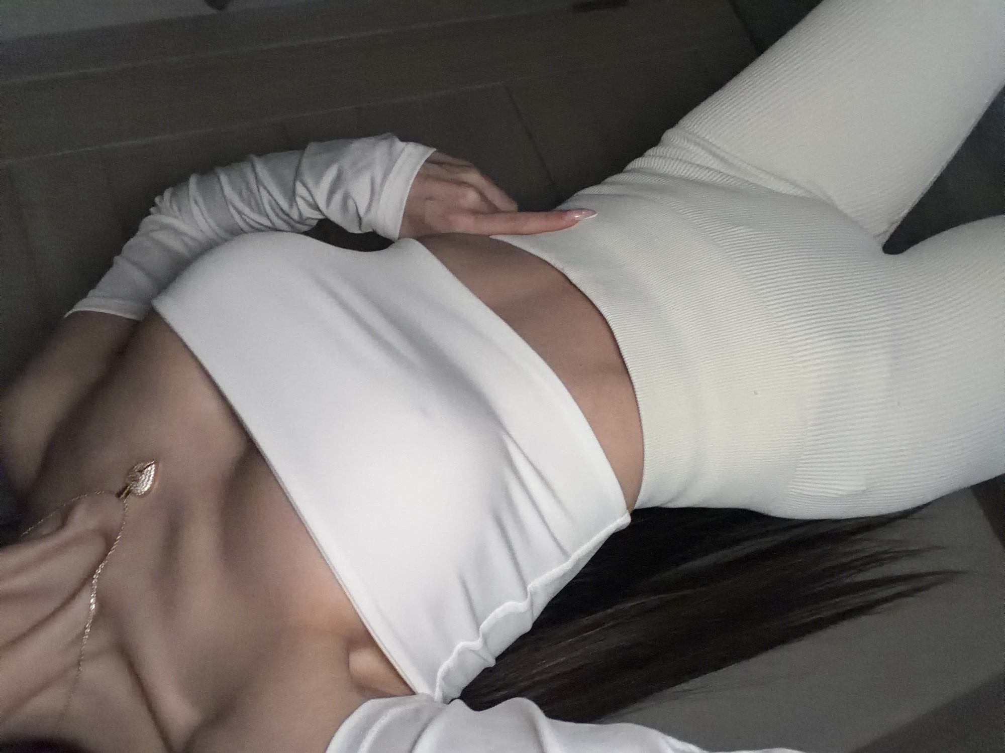 The image captures a person, likely female, lying down indoors, viewed from an oblique, slightly overhead angle. The subject is dressed in form-fitting, light-colored or white activewear, including a long-sleeved crop top that exposes the midriff and ribbed leggings. One arm is raised, with the hand's index finger pointing towards the person's torso. The nails are manicured with a light, possibly pink or nude, polish. A delicate gold-toned chain necklace with a small, leaf-shaped pendant rests on the person's chest. Dark hair is visible fanning out against a dark, featureless background, which could be a floor or a bed. The lighting is soft and even, suggesting an indoor setting. No specific details within the image point to Saint Petersburg, Russia, or any particular time of day. There is no visible text. The overall impression is a posed, casual shot.