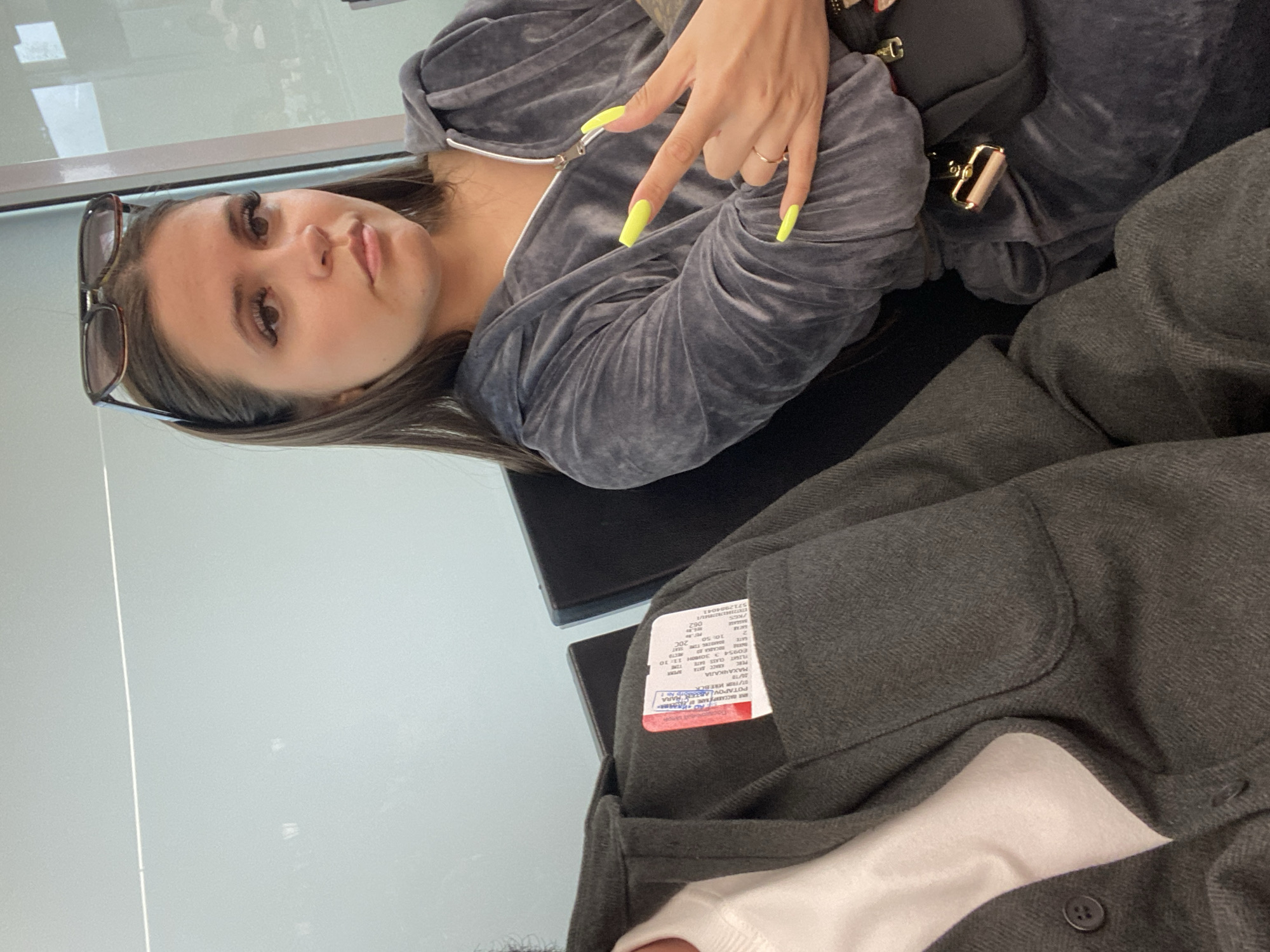 The image shows a close-up of two people sitting in what appears to be an airport waiting area, possibly in Новое Мартьяново, Russia. On the left, a person wearing a dark gray jacket over a white shirt is visible from the chest down. A boarding pass is tucked into the jacket's breast pocket. On the right, a woman with long brown hair, sunglasses perched on her head, and wearing a gray velour tracksuit, is looking towards the camera. Her left hand is raised, displaying bright yellow nails and a peace sign gesture. She has a small black bag with a gold clasp hanging from her shoulder. The background consists of muted tones, with hints of white seating and large windows.