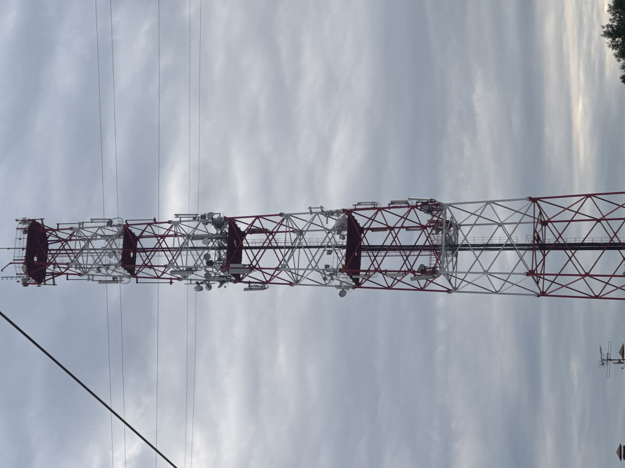 The image shows a tall, red and white lattice telecommunications tower against a cloudy sky. The tower is equipped with numerous antennas and satellite dishes at various levels. Several horizontal power lines are visible crisscrossing in front of the tower and the sky. In the lower left portion of the image, a small portion of another structure with a peaked roof and some antennas is visible, along with the tops of some trees on the right. The scene appears to be an outdoor, industrial or infrastructure area.