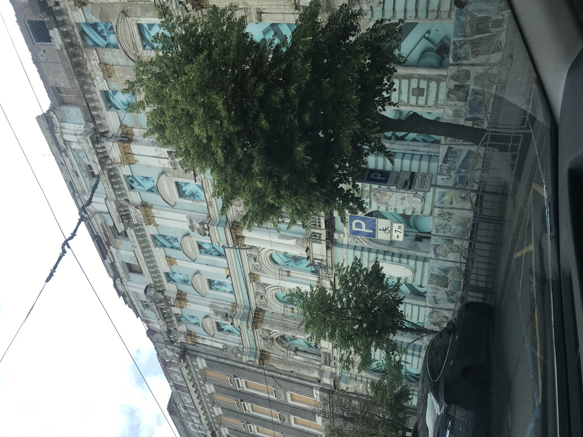 Captured from inside a vehicle, the image shows a multi-story building in Kharkiv, Ukraine. The building's architecture is characterized by classical elements, including ornate facades, columns, and detailed window frames. However, the exterior appears worn and in need of renovation, with visible signs of decay and discoloration. Some sections of the building feature light blue accents. The view of the building is partially obscured by trees, which are situated along the street. Parking signs are visible next to the building. A dark-colored car is partially visible in the foreground. The shot is taken at an oblique angle, with the building appearing tilted. The sky is overcast, indicating a cloudy day.