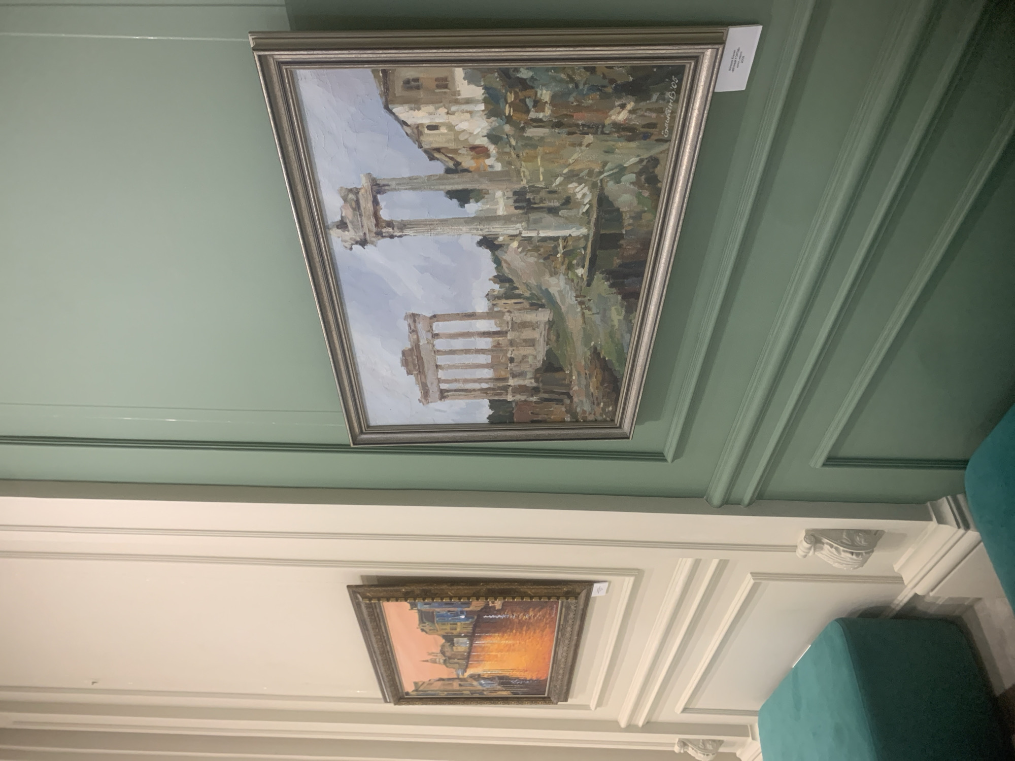 The image shows a section of a room in Izhevsk, Russia. Two framed paintings hang on a pale green and white paneled wall. The top painting depicts ancient Roman ruins in a painterly style, while the bottom painting shows a cityscape with a sunset. A teal upholstered bench is visible in the lower right corner.