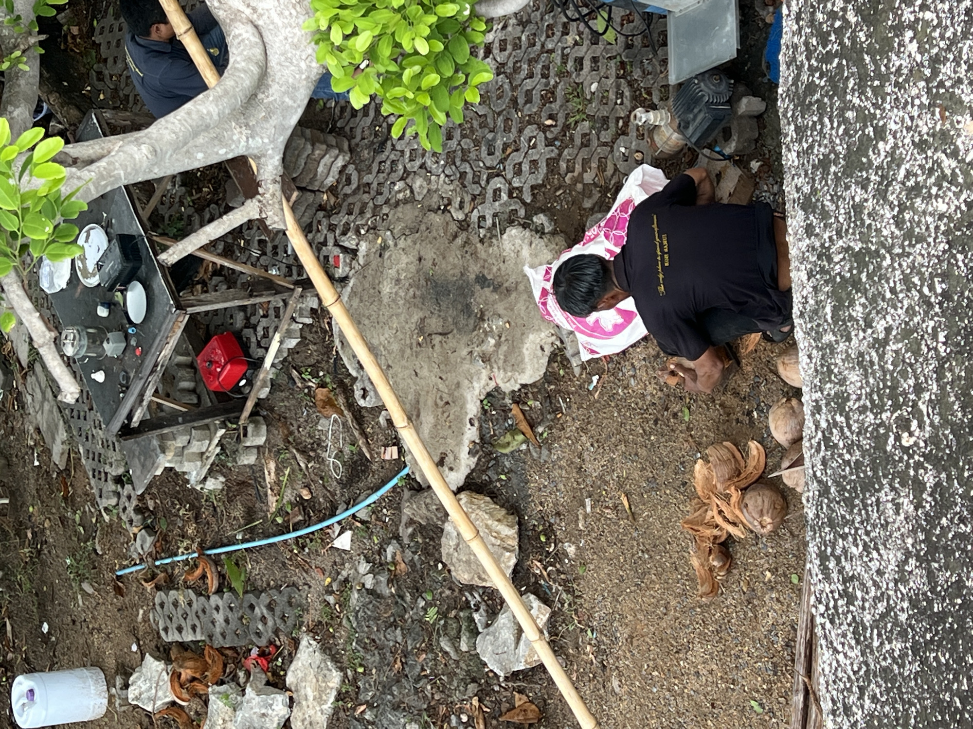 The scene is an outdoor area in Baan Chaweng Noi, Thailand. A man wearing a black t-shirt is sitting on a patterned mat and working with coconuts near a textured grey wall. Scattered around him are several whole coconuts and coconut husks. Nearby, there is a section of patterned grey pavers, a patch of greenery, and a makeshift table with various objects, including white plates, a red canister, and other unidentified equipment. A long bamboo pole stretches across the ground from near the table to the area where the man is working. A blue pipe and a white container also lie on the ground. Another person is partially visible seated on a small stool, near the table. The ground surface is a mix of earth, gravel, and scattered debris.