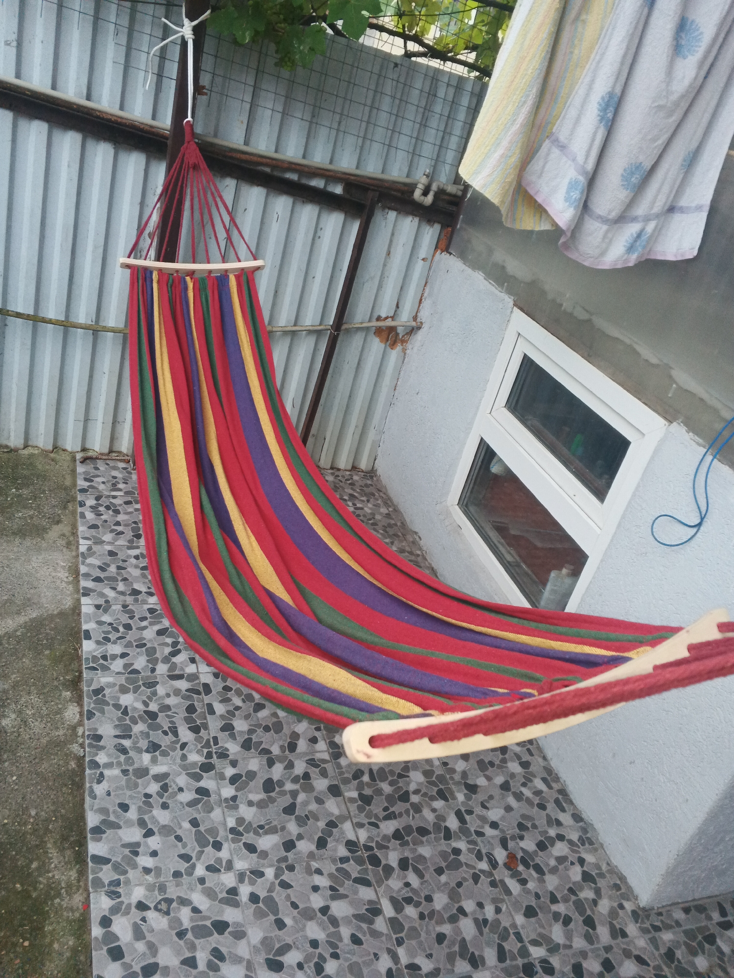 A colorful striped hammock hangs outdoors, suspended from a metal pole. The hammock is made of fabric with red, yellow, green, and purple stripes and has wooden spreaders at each end. It is positioned in front of a corrugated metal fence and a light-colored building with a small white-framed window. Two towels are hanging on a line above the fence, one with a yellow and white stripe pattern and the other a light blue with a floral pattern. The ground is covered with grey and black mosaic tiles. The scene appears to be in a backyard or patio area. There is no visible text. The lighting suggests it is daytime.