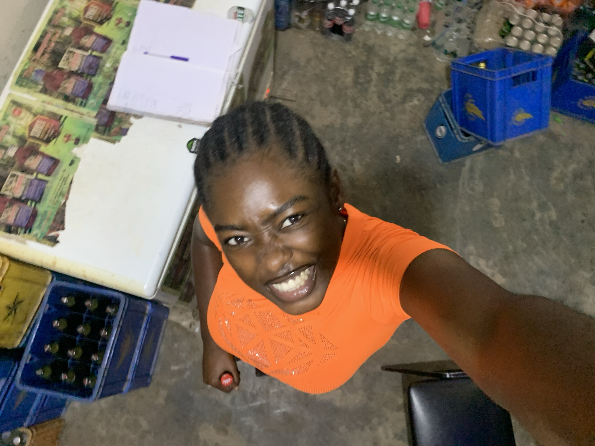 A high-angle selfie depicts a Black woman with cornrow braids, wearing an orange shirt. She has a broad smile.
The setting is a possibly indoor concrete floor. A table with papers is visible to the left of the frame. Several blue crates, some containing bottles, are nearby. A blue crate is also visible. In the background are numerous bottles and cans.
She is holding a bottle cap in her right hand.
It is not possible to verify the location within the city of Kabong, Nigeria from the images provided.